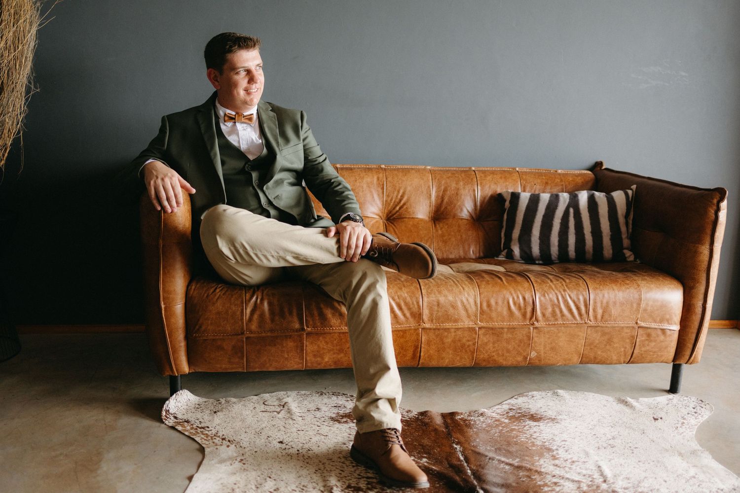 Someone sits casually on a tan leather sofa with striped pillows against a dark wall in a modern interior setting.