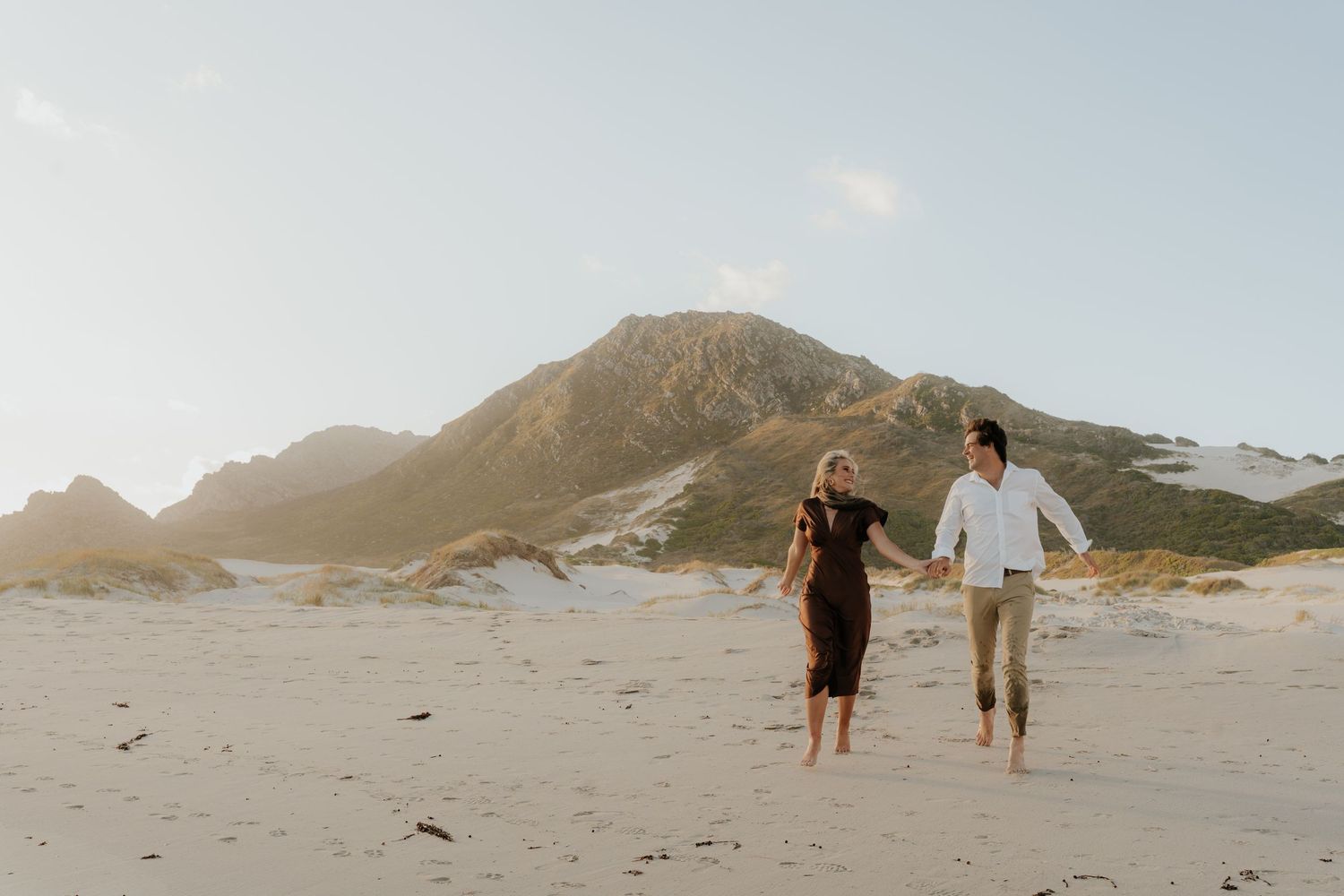 A couple holds hands and runs joyfully on a sandy beach with a mountain backdrop in warm sunlight.