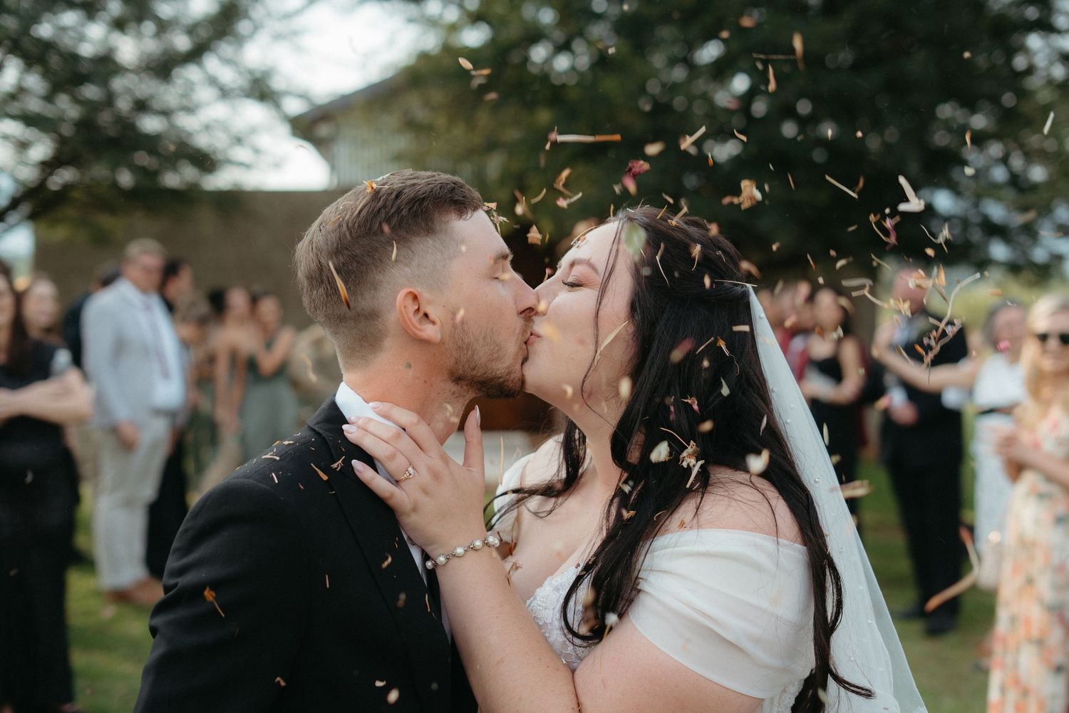A romantic kiss between newlyweds as confetti falls around them during an outdoor celebration.