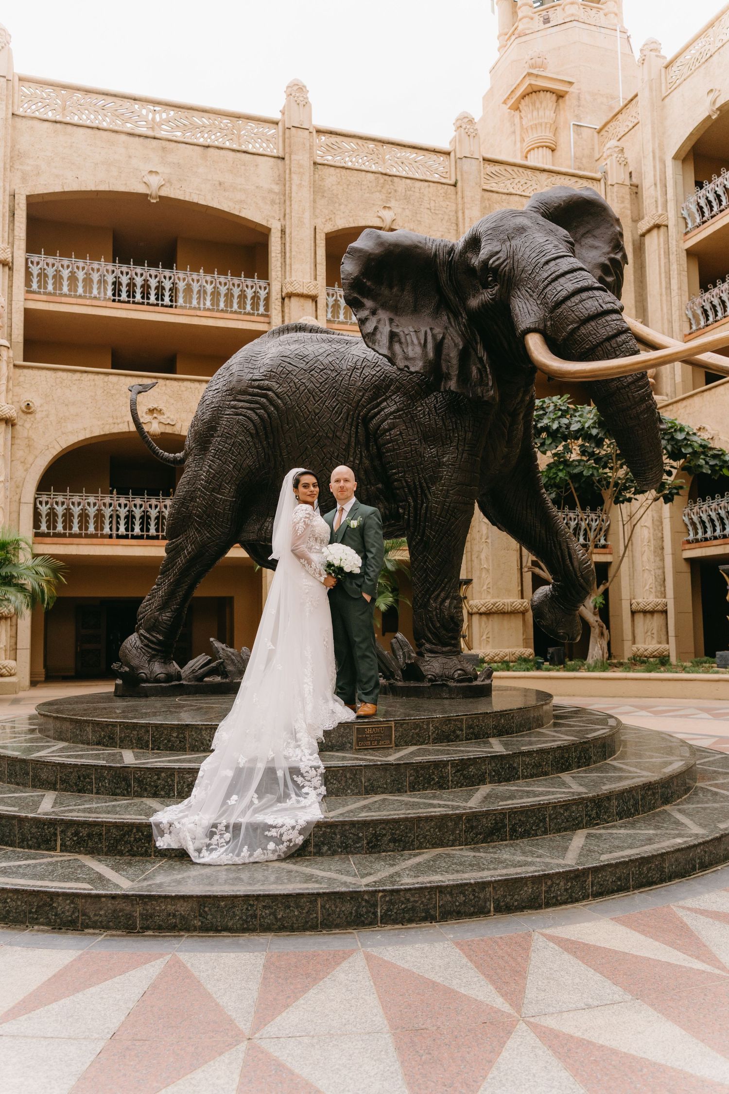 Wedding couple poses in front of large stone elephant statue on a circular platform at ornate colonial-style building.