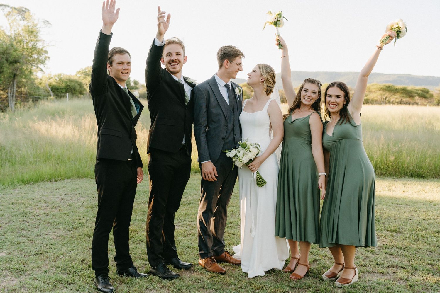 Wedding party posing in a field with bridesmaids in green dresses and groomsmen in black suits.