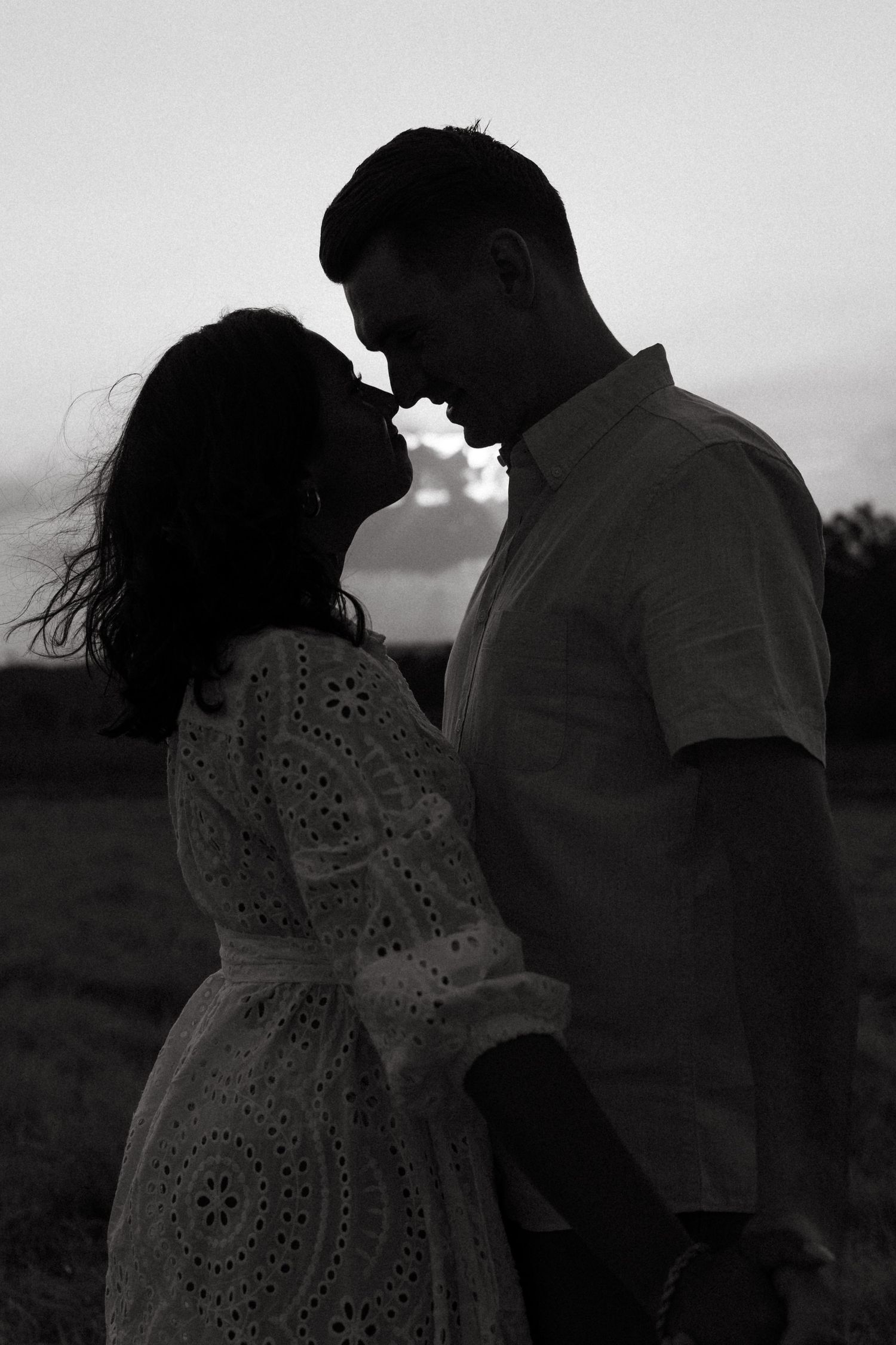Silhouetted couple shares intimate moment in field during dramatic black and white sunset portrait session.