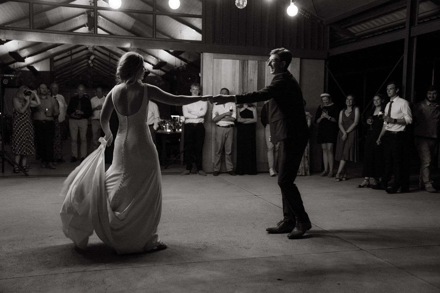 A black and white photo captures newlyweds during their first dance in a rustic venue with string lights.