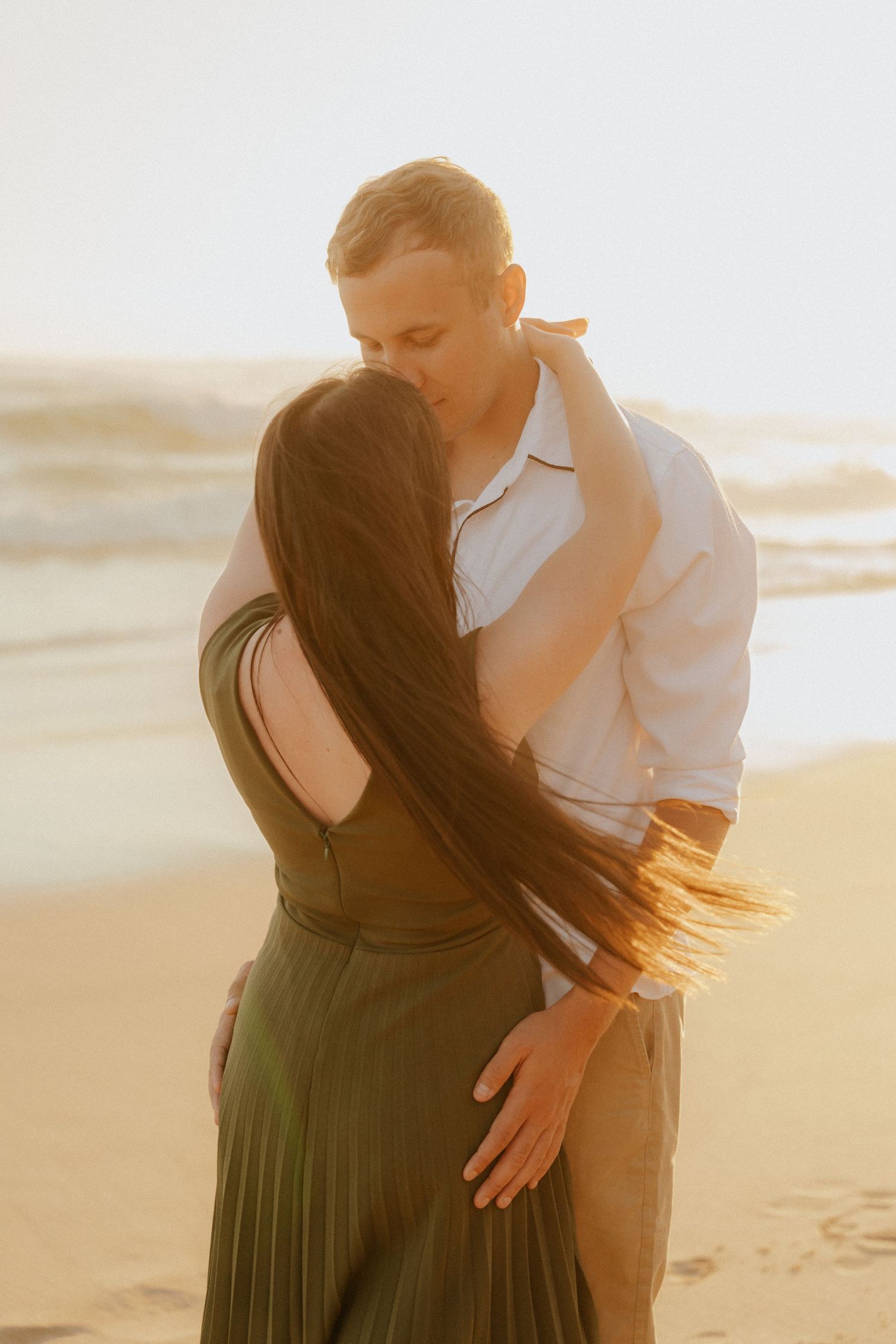 A couple embraces romantically on a beach at sunset, her dark dress flowing in the golden light.