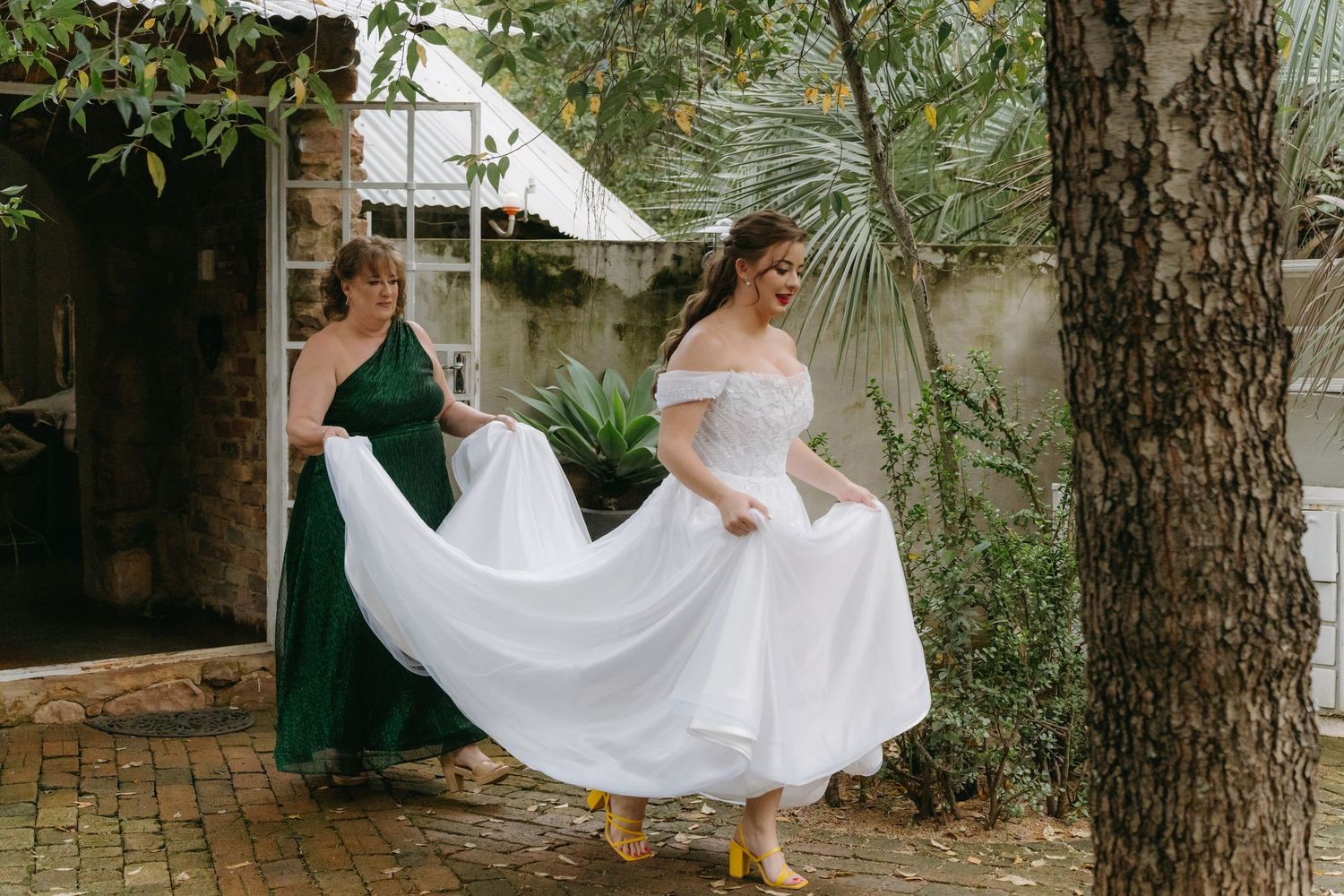 Wedding preparations with flowing white dress being arranged outdoors among trees.