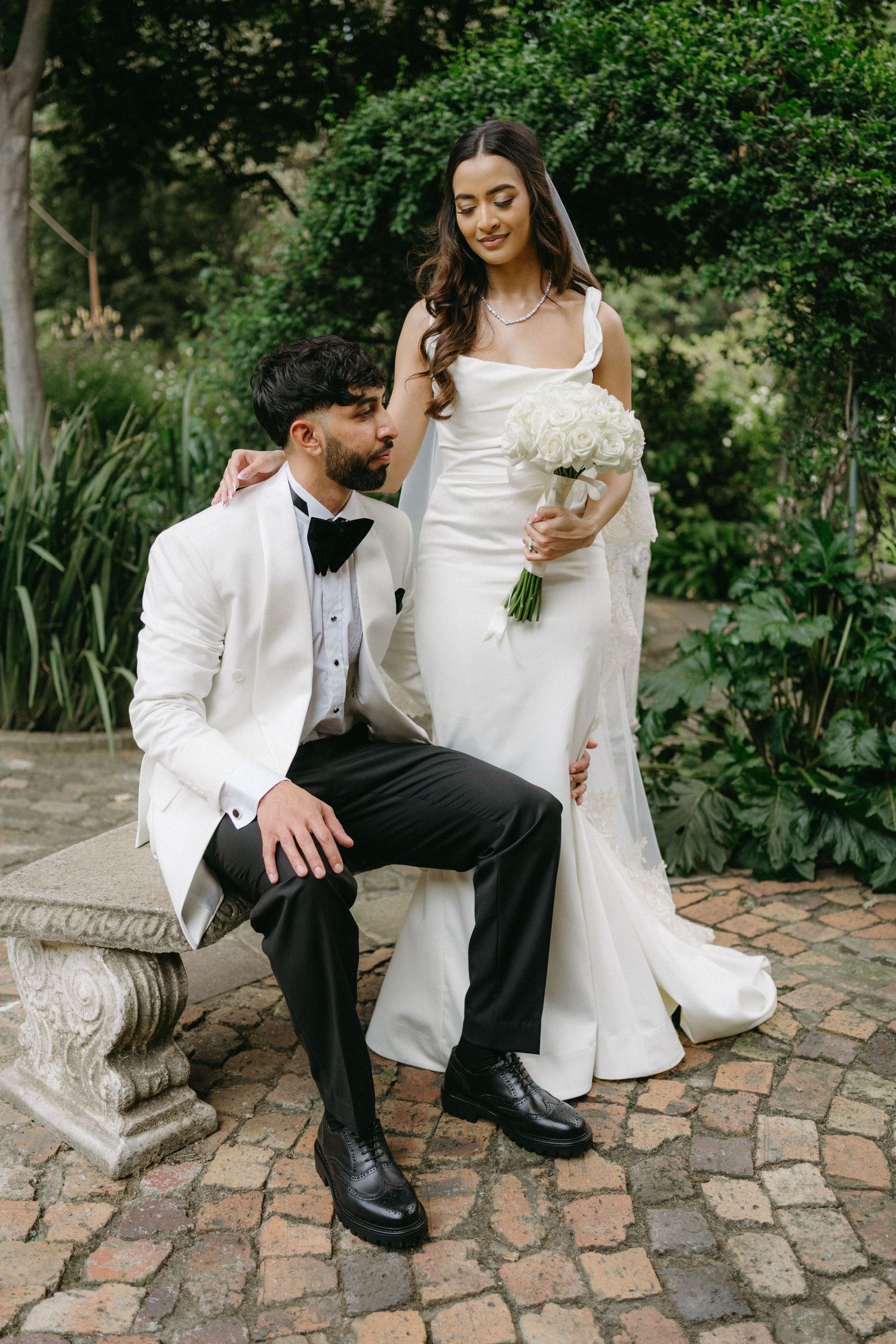 Elegant wedding couple poses on stone bench in garden setting with bride holding white bouquet.