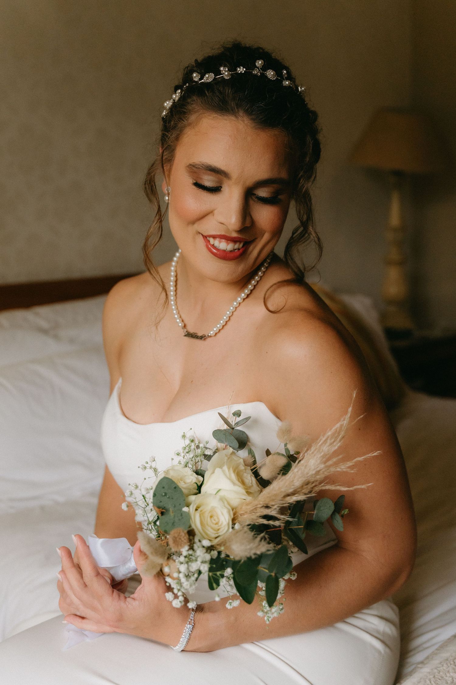 A bride in a white dress sits on a bed holding an elegant bouquet of white roses and eucalyptus.