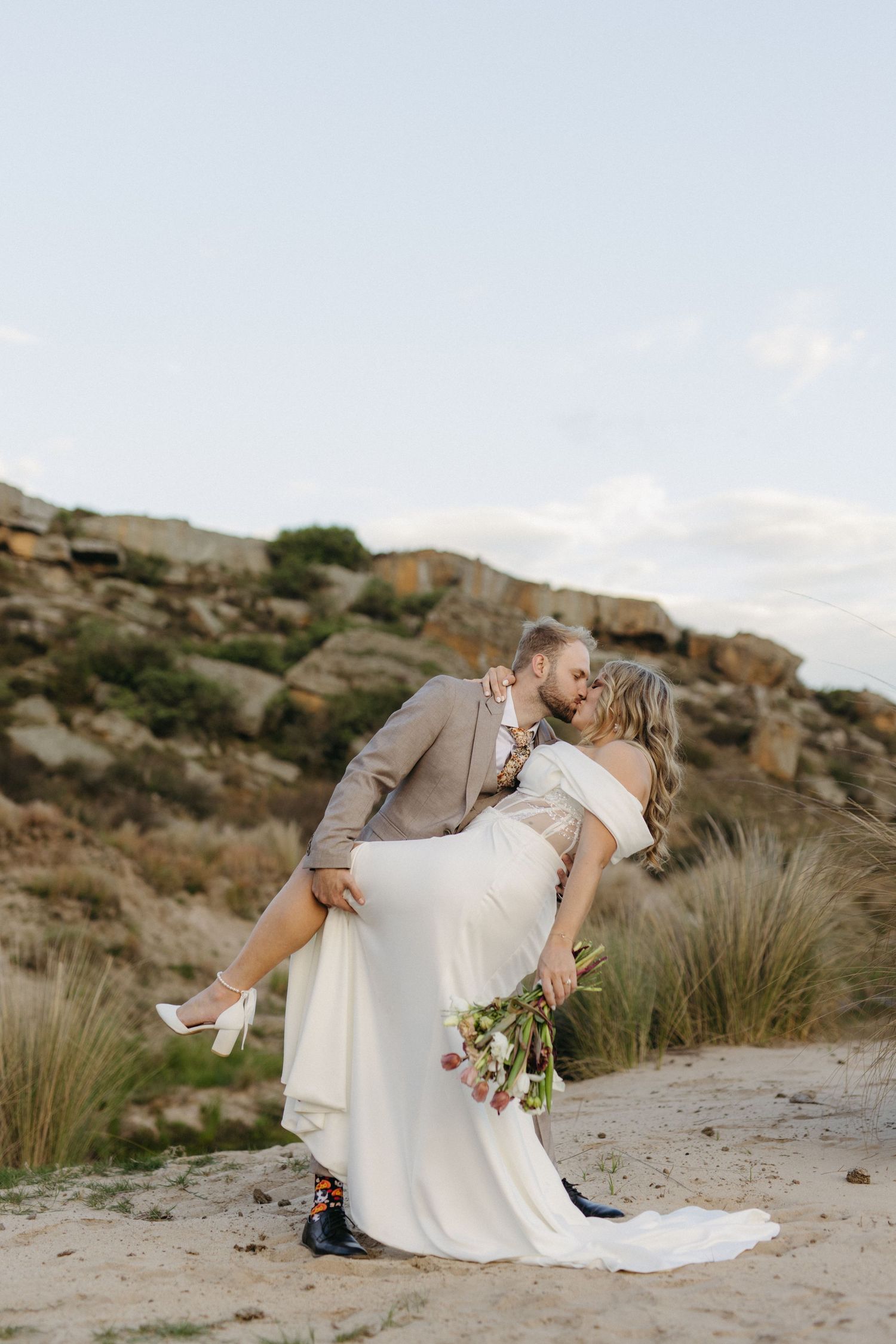 Couple shares romantic dip kiss pose against rocky desert landscape at sunset while bride wears flowing white dress.