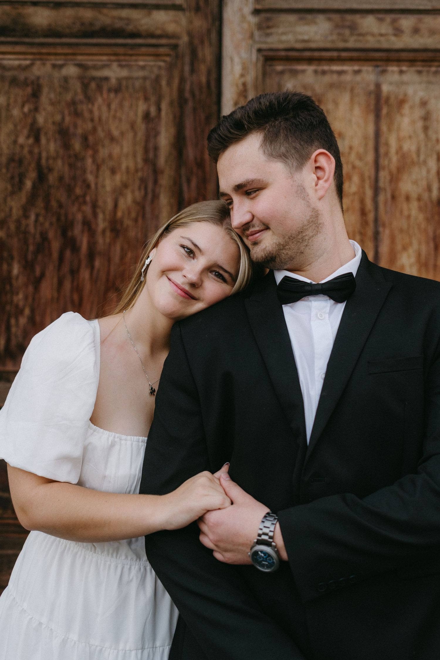 A romantic wedding couple embraces against rustic wooden doors, with the bride in white and groom in classic tuxedo.