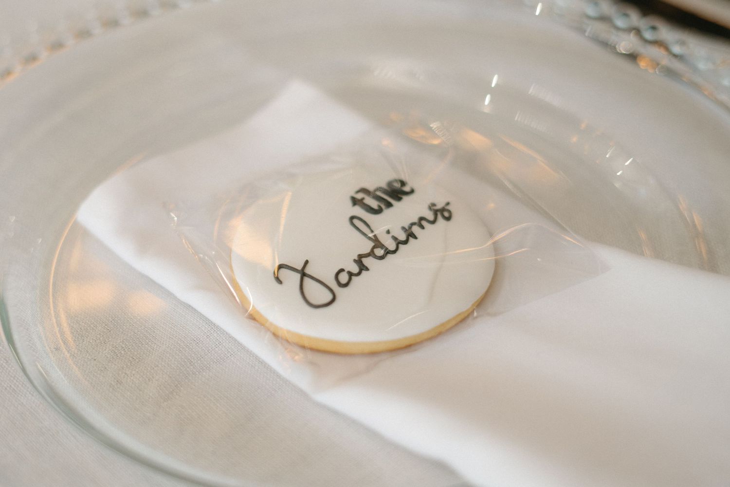 A sugar cookie with 'the Jardims' handwritten text sits on a white napkin on a glass plate.