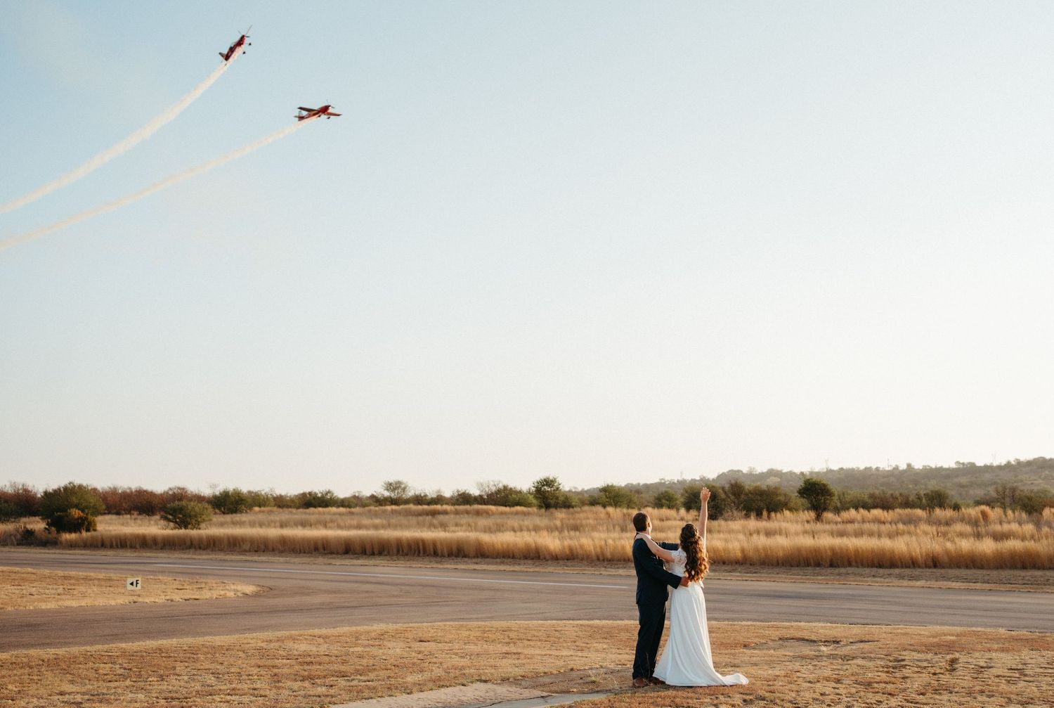 Two airplanes flying overhead while a couple stands in a romantic pose on an empty runway at sunset.