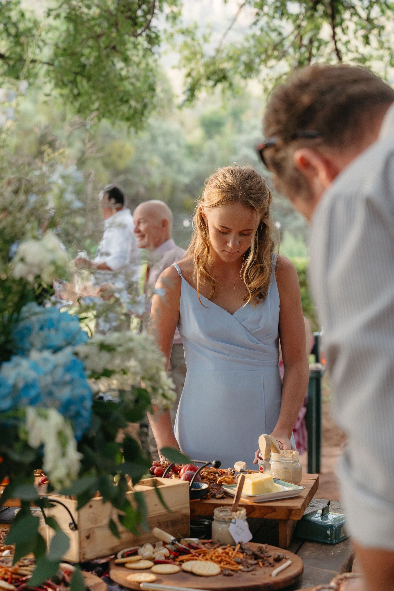 Wedding guests gather around a rustic outdoor dining table with appetizers under tree branches.
