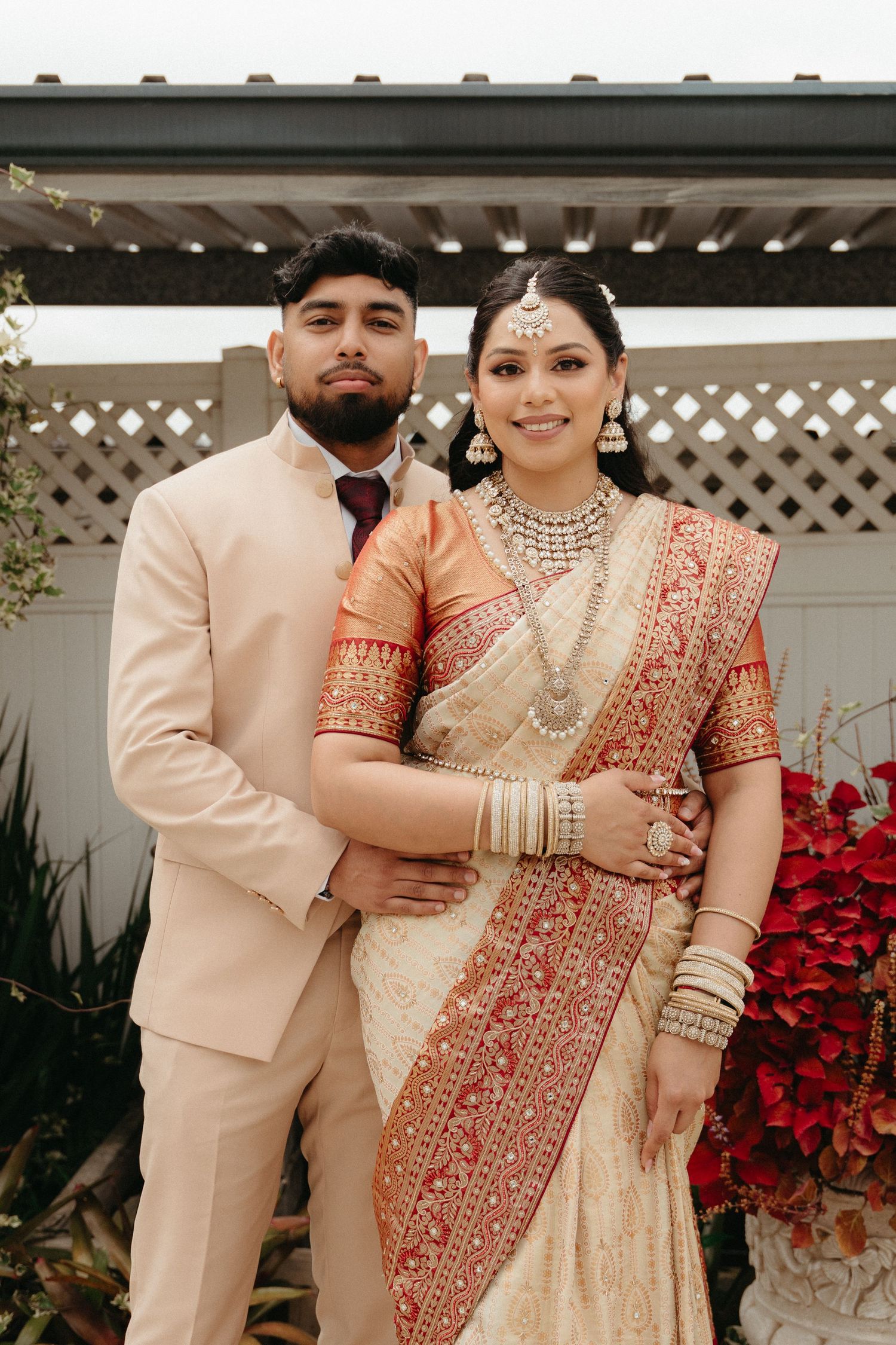 A couple in traditional Indian wedding attire pose together against a white lattice backdrop with red poinsettias.