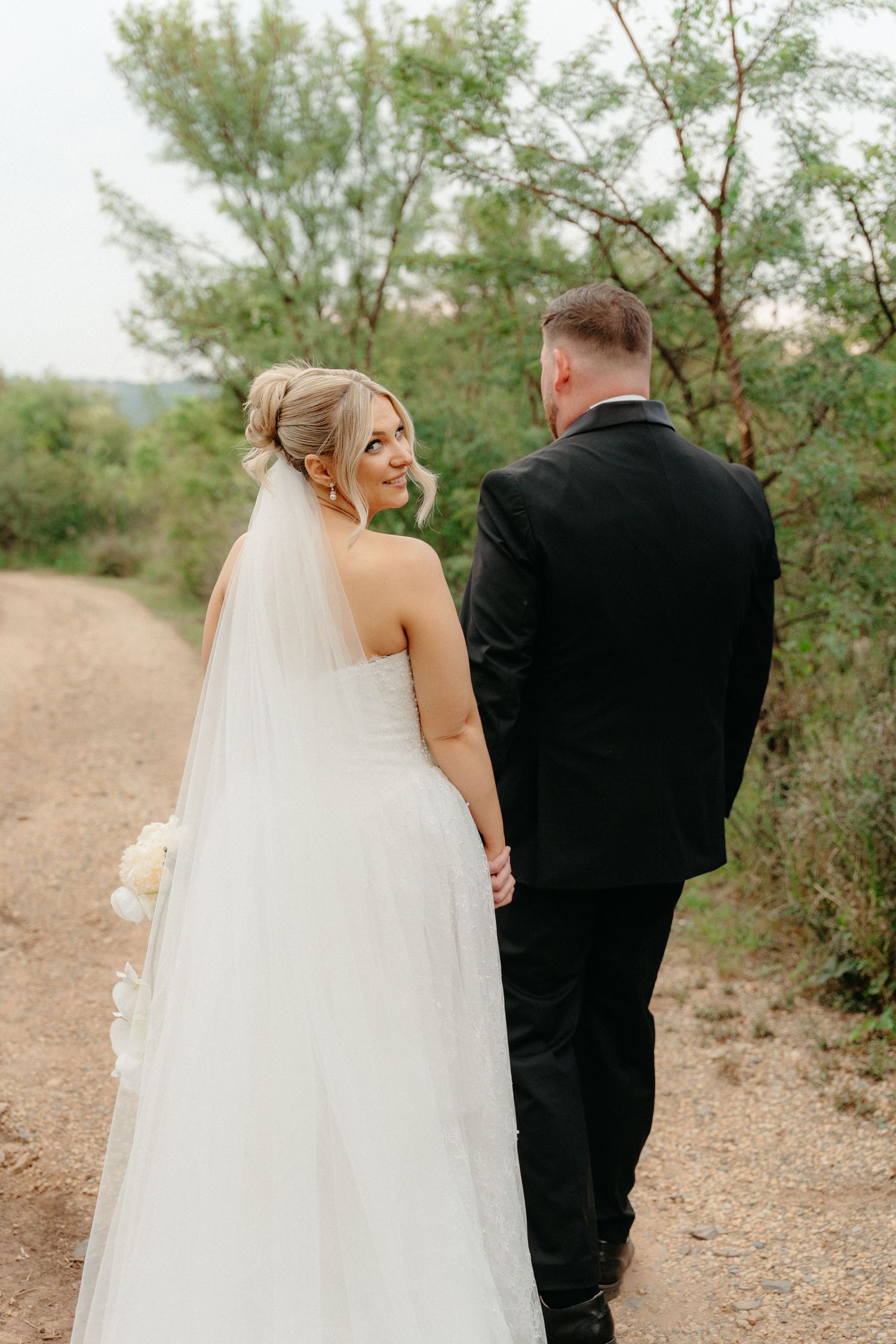 A couple walks along a desert path in wedding attire with a long flowing veil trailing behind them.