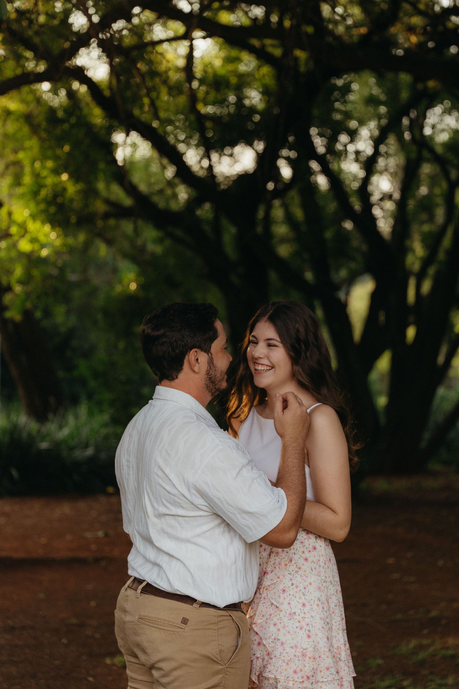 A couple shares a tender moment outdoors, smiling at each other beneath lush green trees in warm golden light.