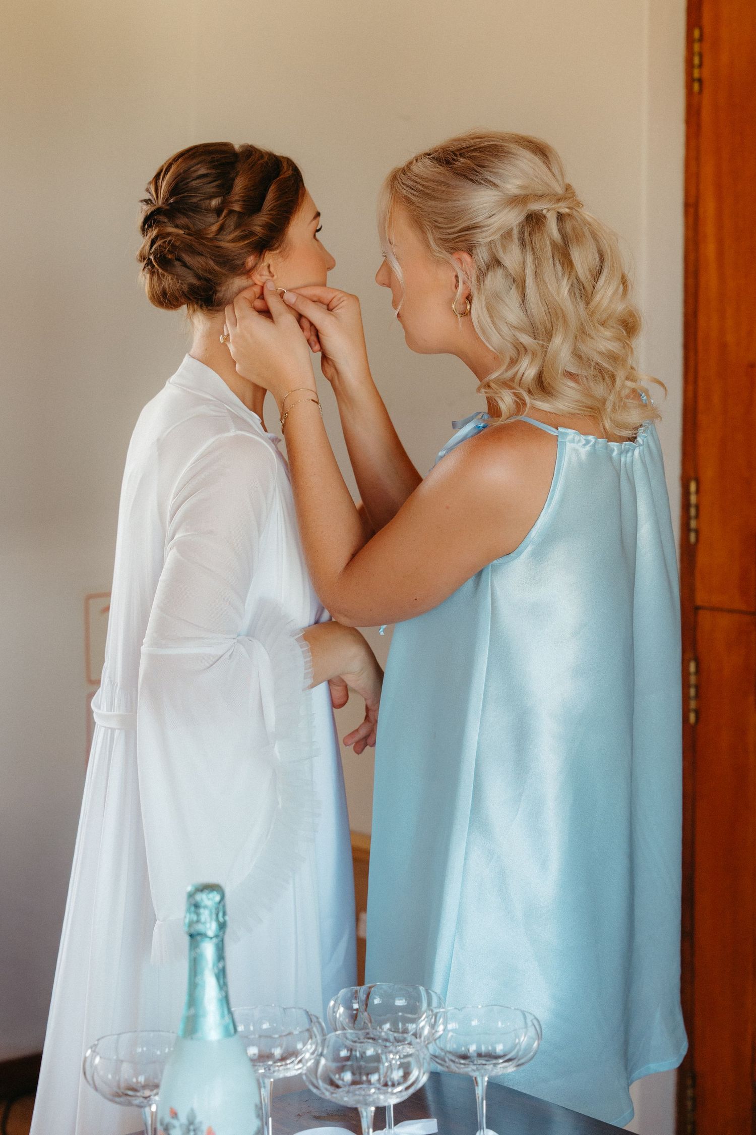 Two people in elegant attire help each other get ready near a table with champagne glasses and a blue bottle.