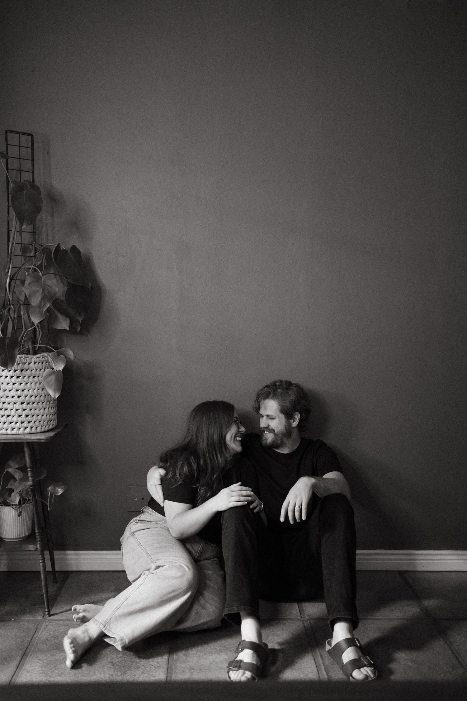 Black and white photo of a couple sitting on the floor together in Pretoria, smiling and embracing near a houseplant.
