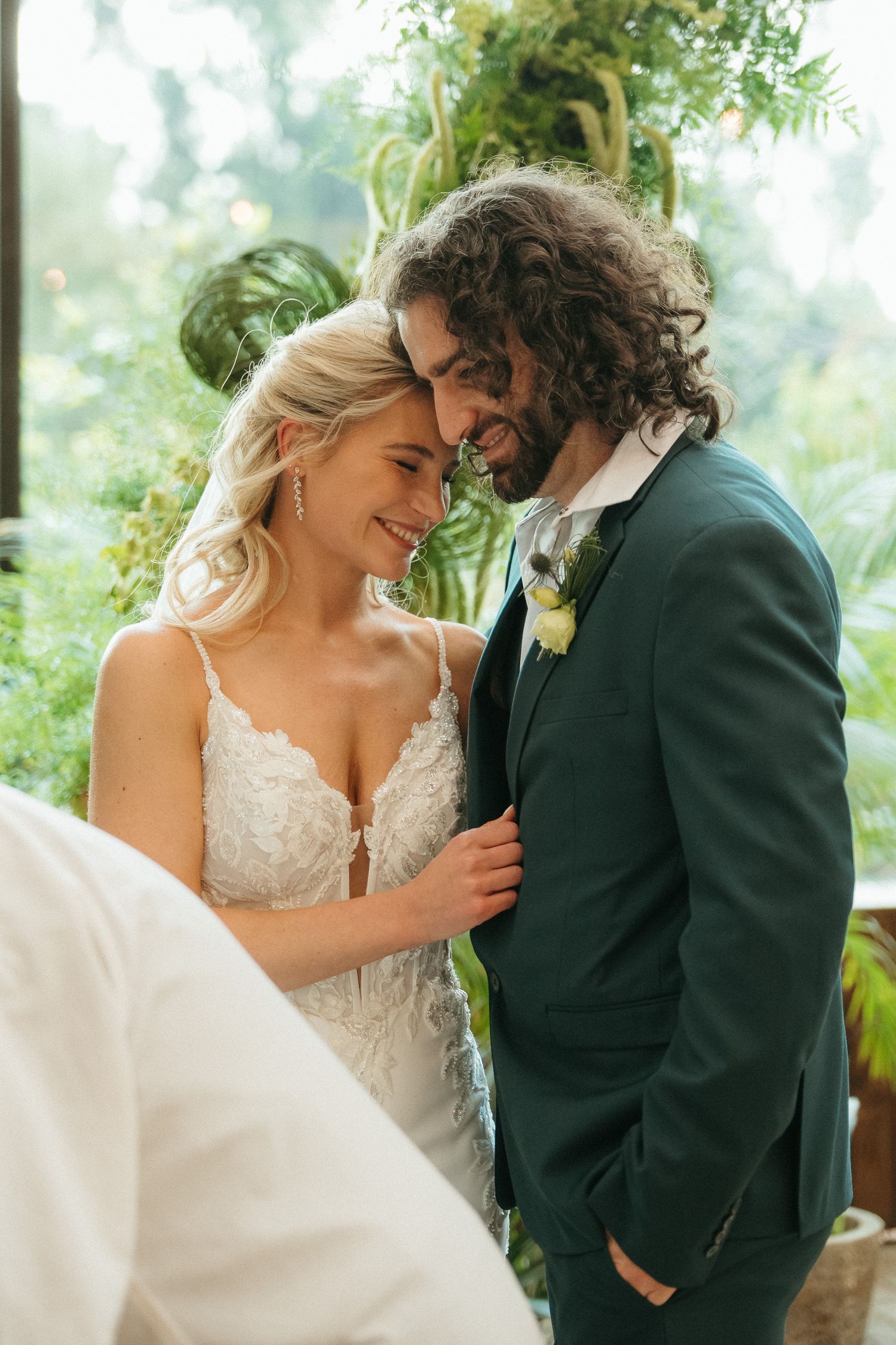 A bride and groom share an intimate moment, smiling together in a lush green botanical setting on their wedding day.
