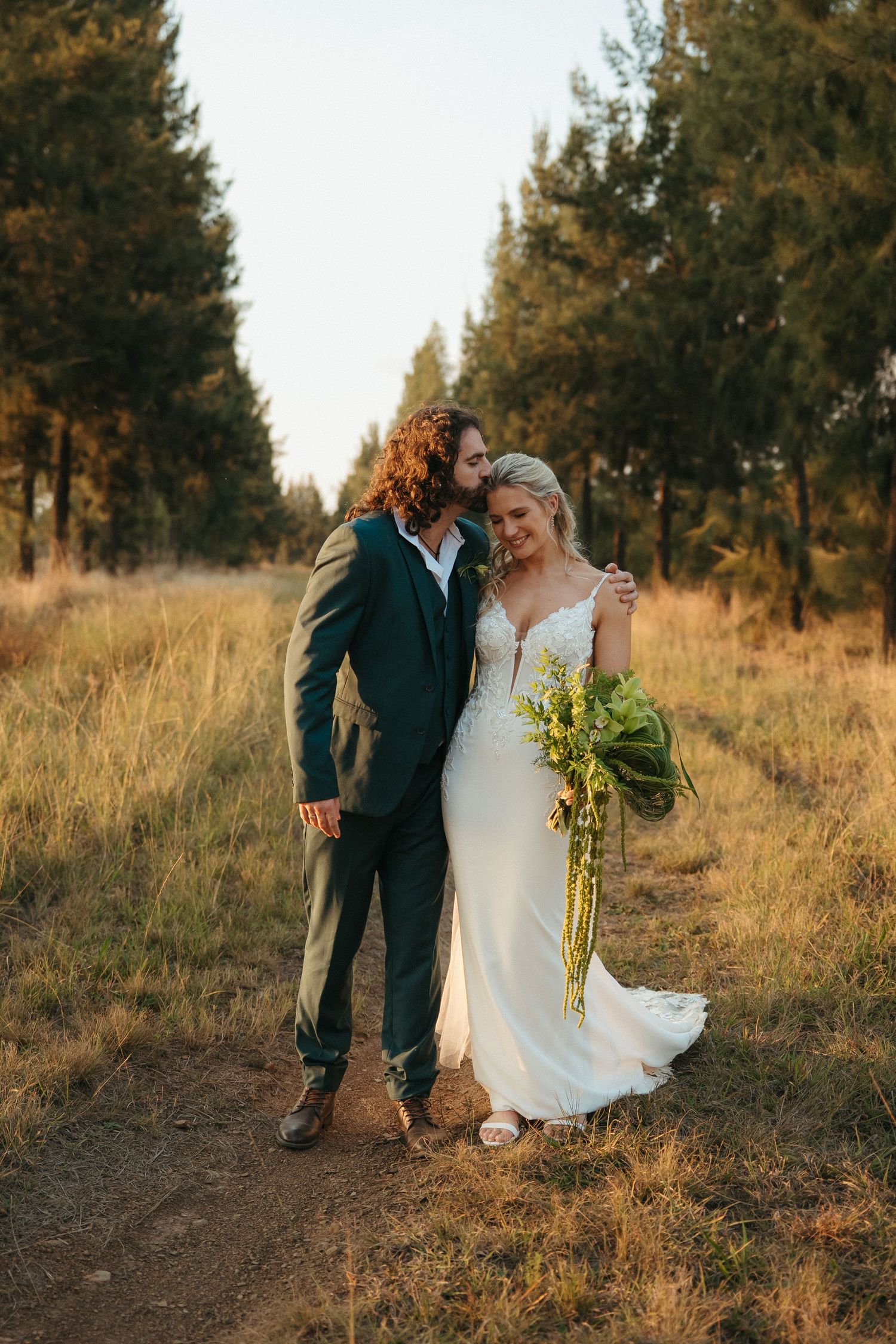 Bride and groom embracing on a golden field path surrounded by tall pine trees during sunset wedding portrait.