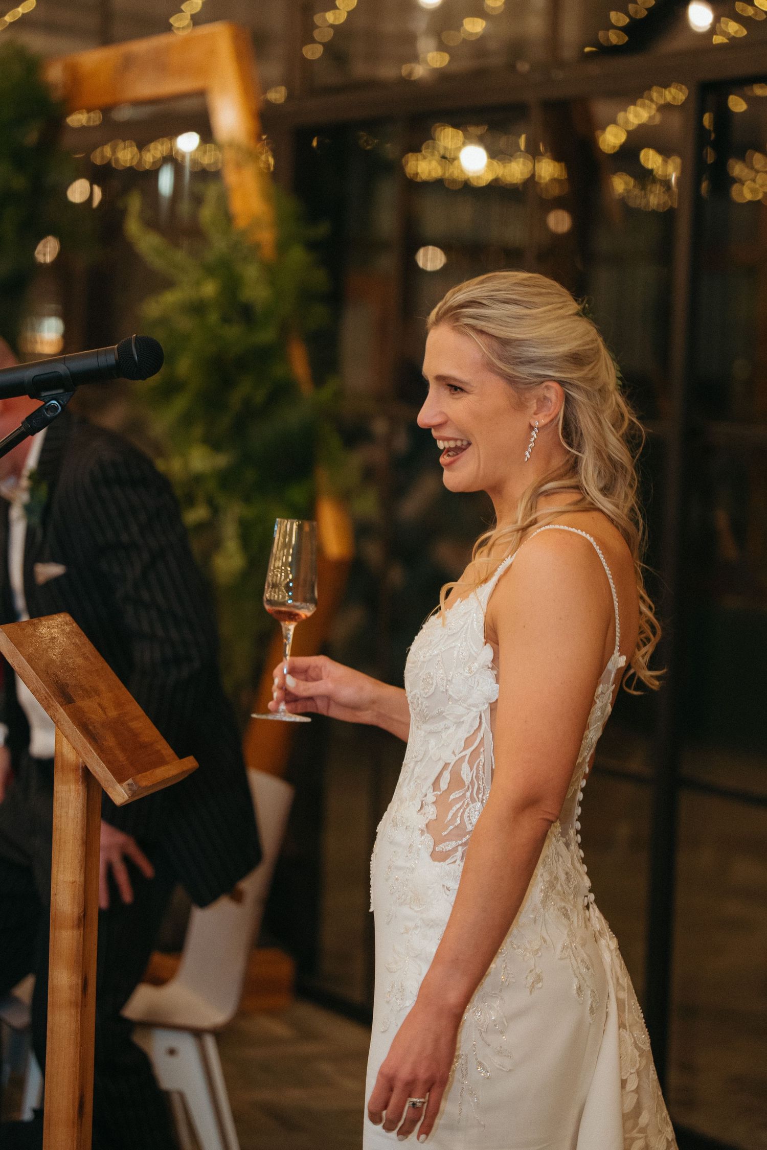 Smiling bride in lace wedding gown holding champagne glass during reception toast with fairy lights backdrop.