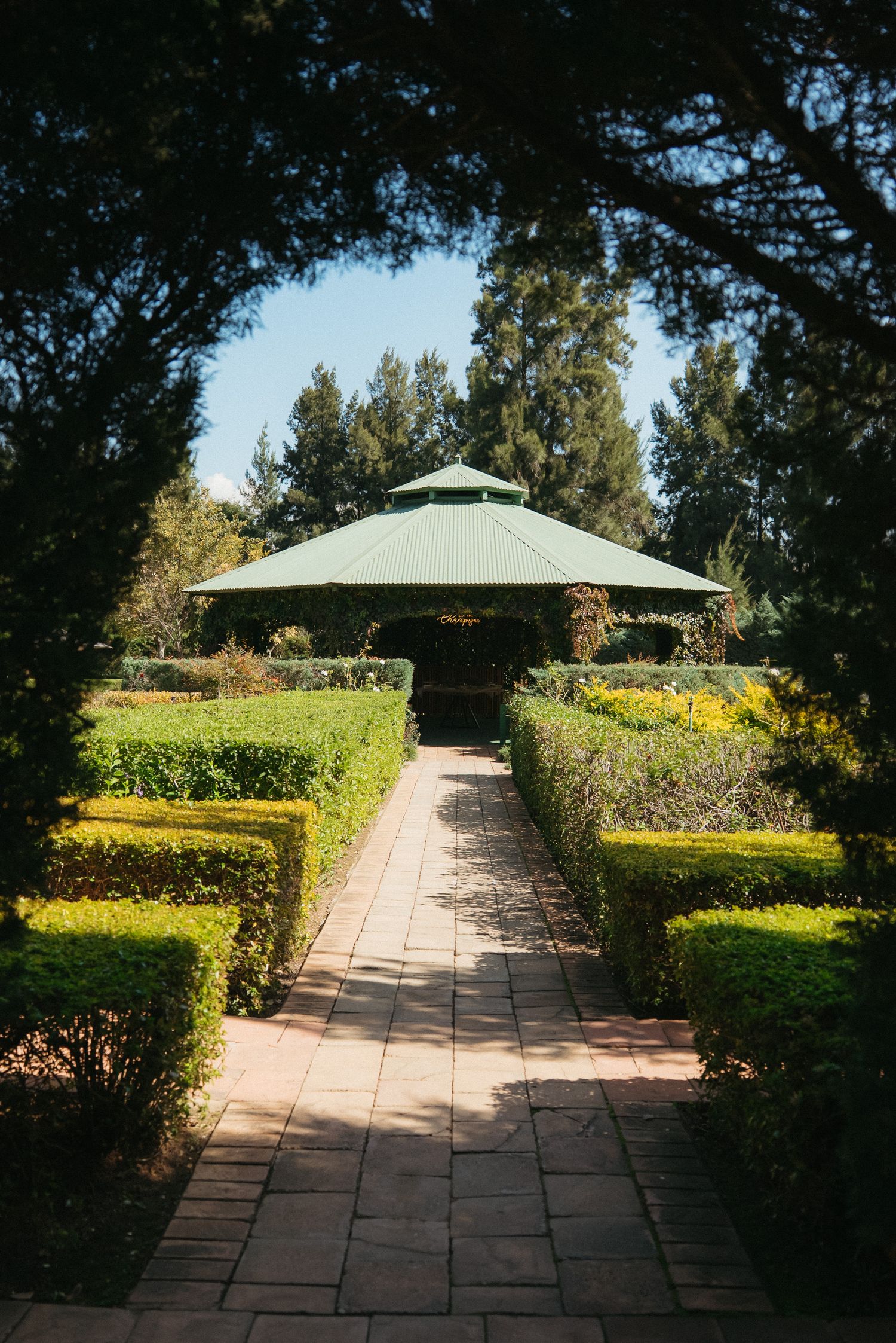 Brick pathway leading to a green gazebo surrounded by manicured hedges and tall trees in a formal garden.