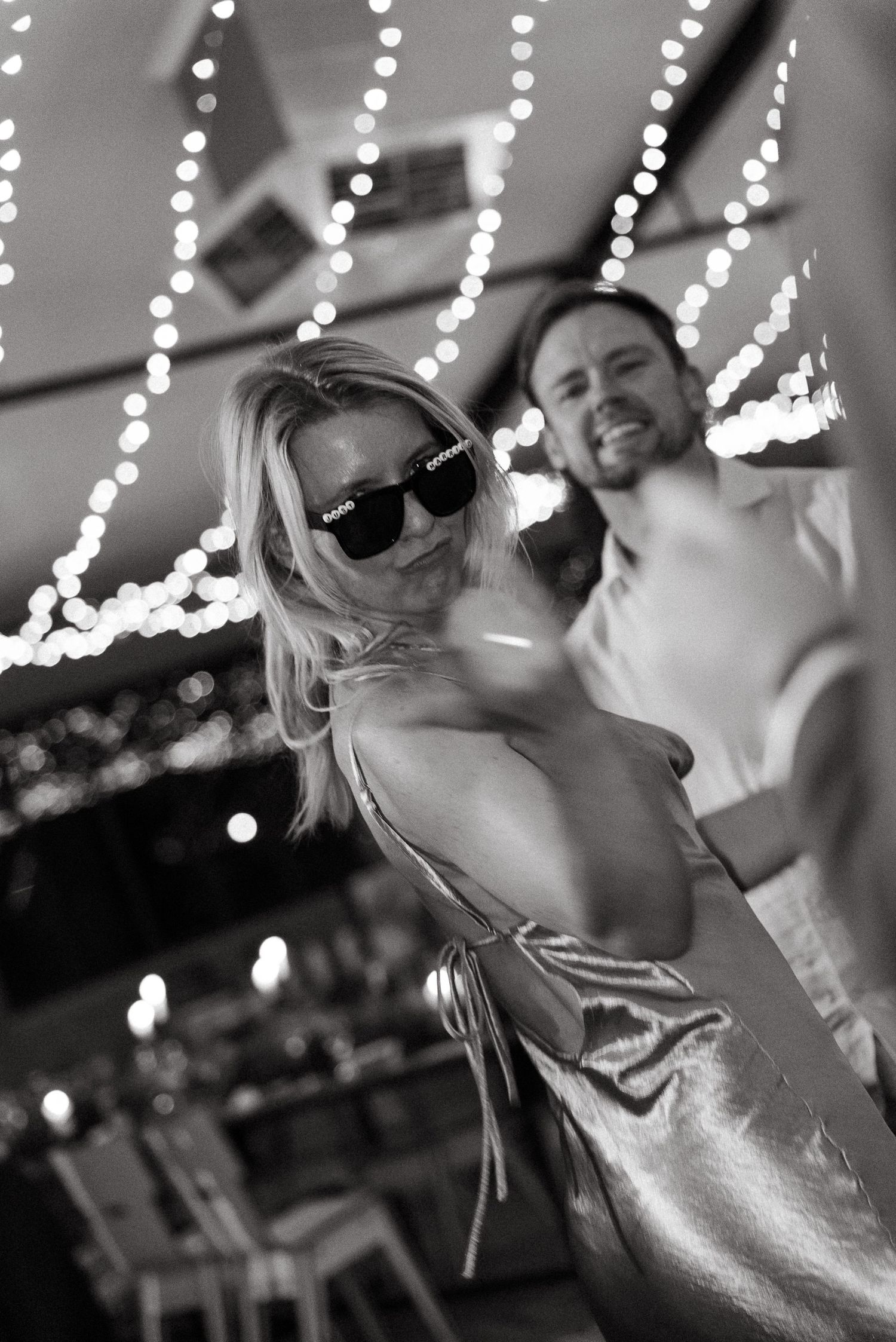 Black and white photo of a blonde woman in sunglasses and a man smiling under string lights at a festive event.