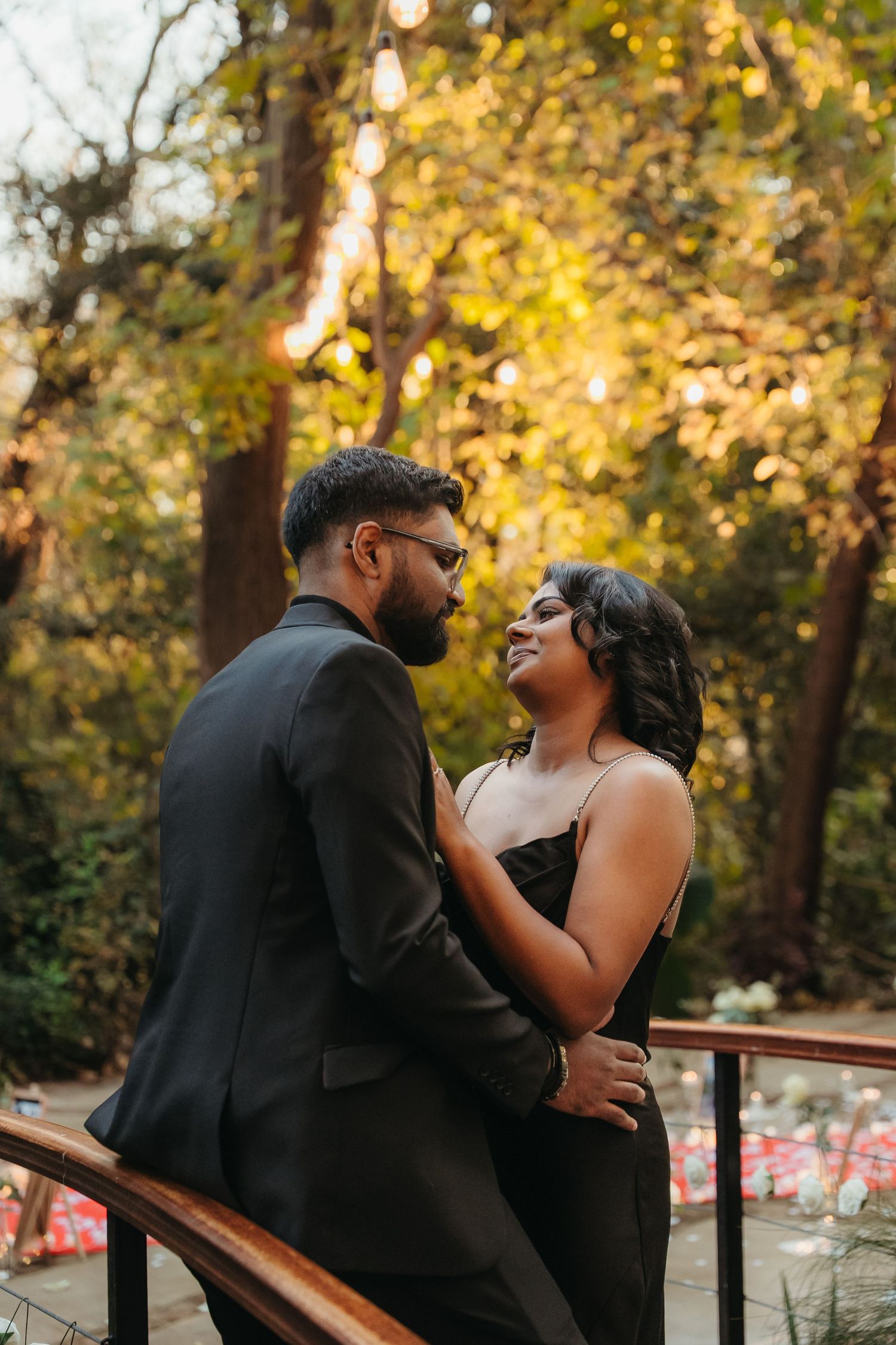 Couple in black outfits sharing a romantic moment on a balcony surrounded by golden autumn trees at dusk.