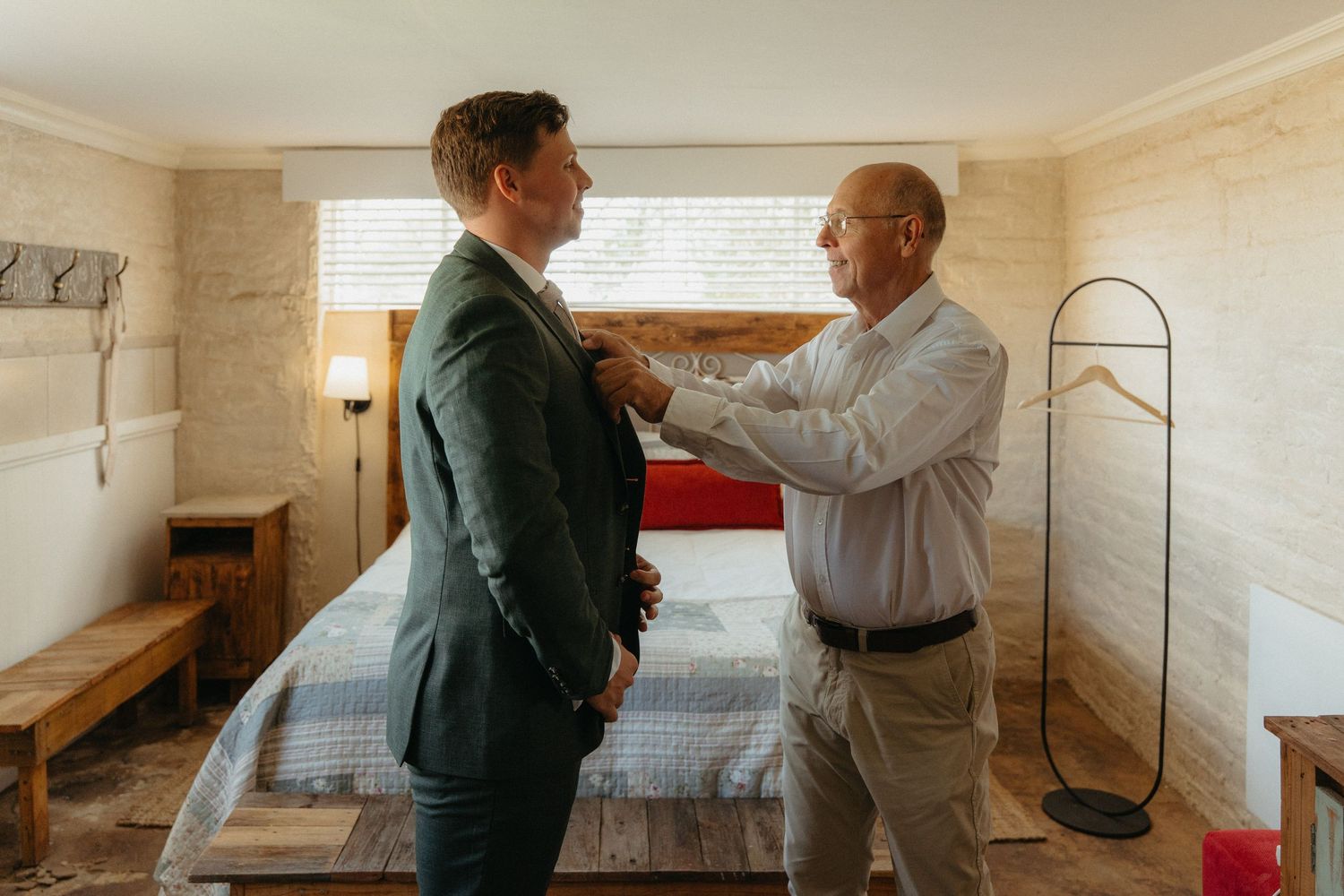 Two men in a bedroom, one adjusting the other's suit jacket, preparing for a formal event.