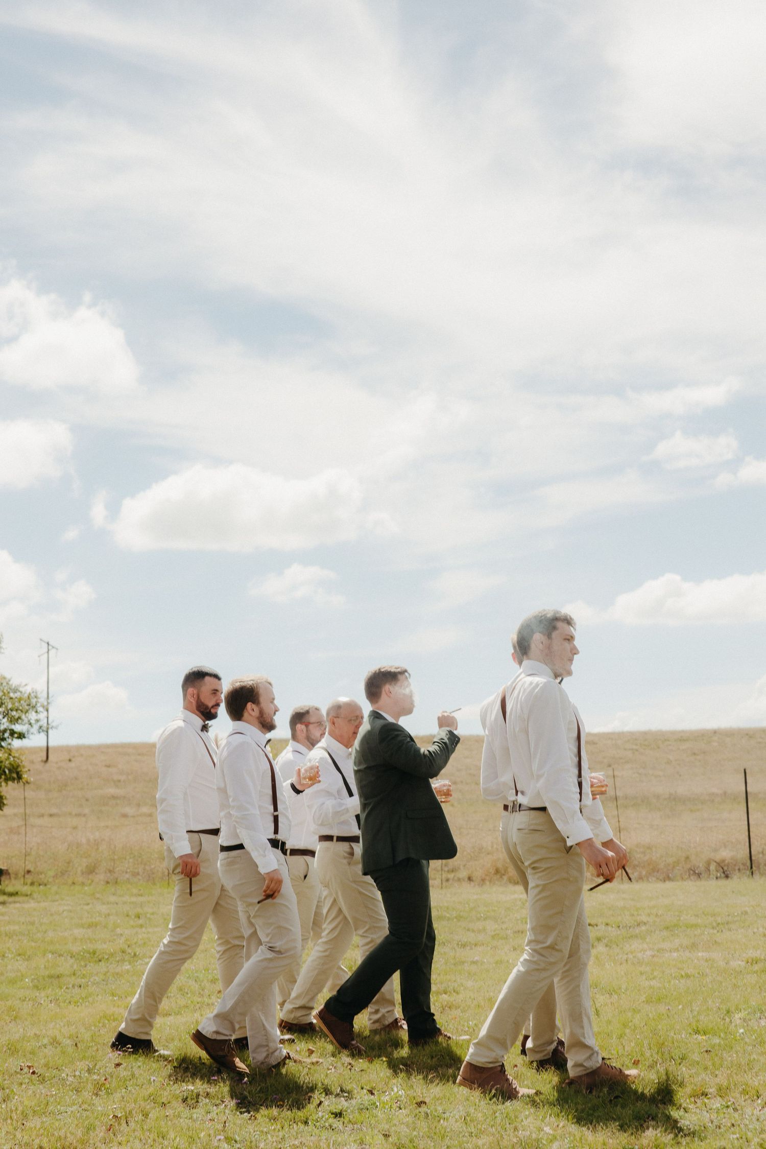 Groom in dark suit walks with groomsmen in white shirts and khaki pants across a sunny green field.