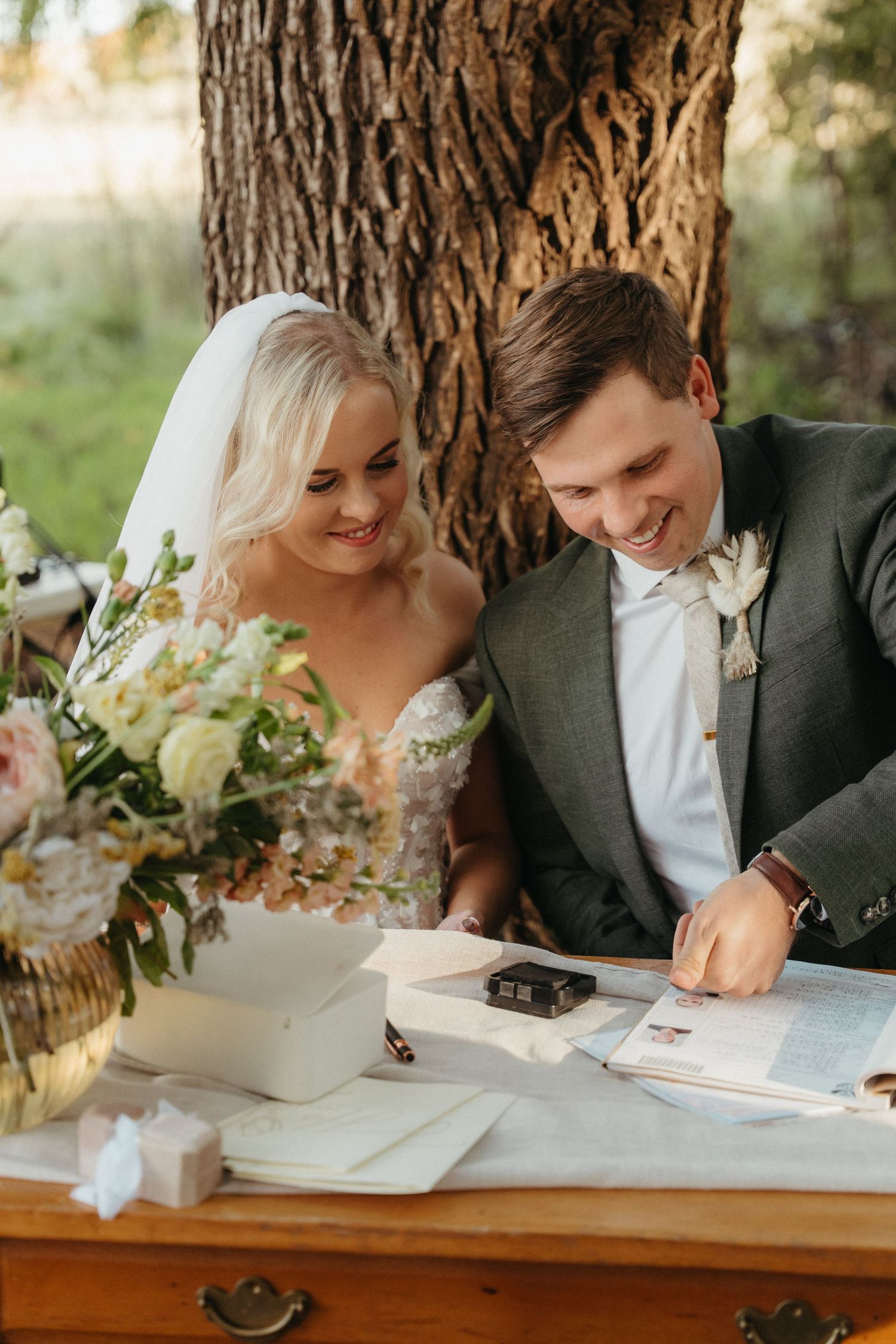 Smiling bride and groom signing wedding registry documents at wooden table with floral bouquet outdoors near large tree.