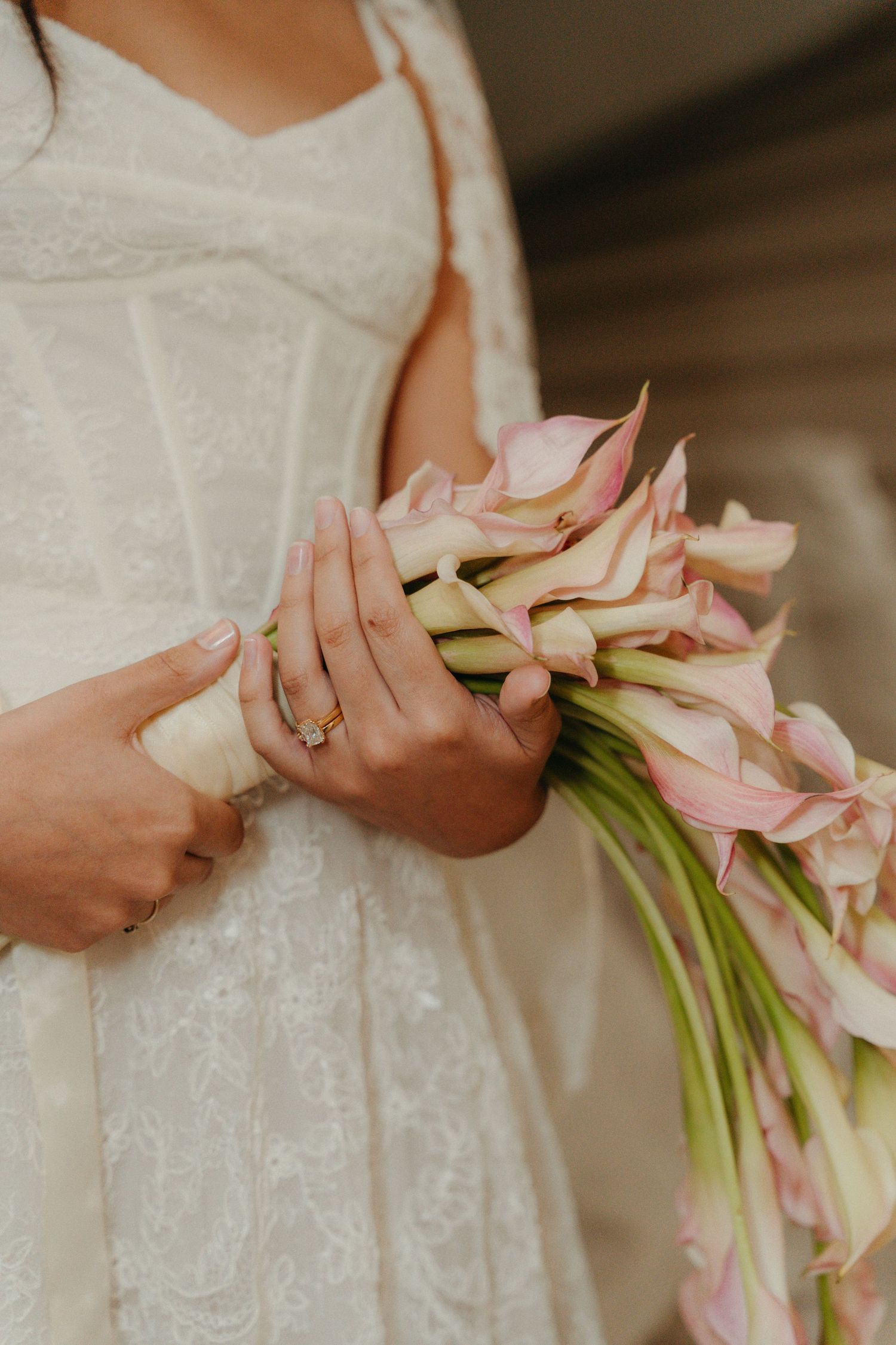 Bride in white lace dress holding pink calla lily bouquet with diamond engagement ring visible on her hand.
