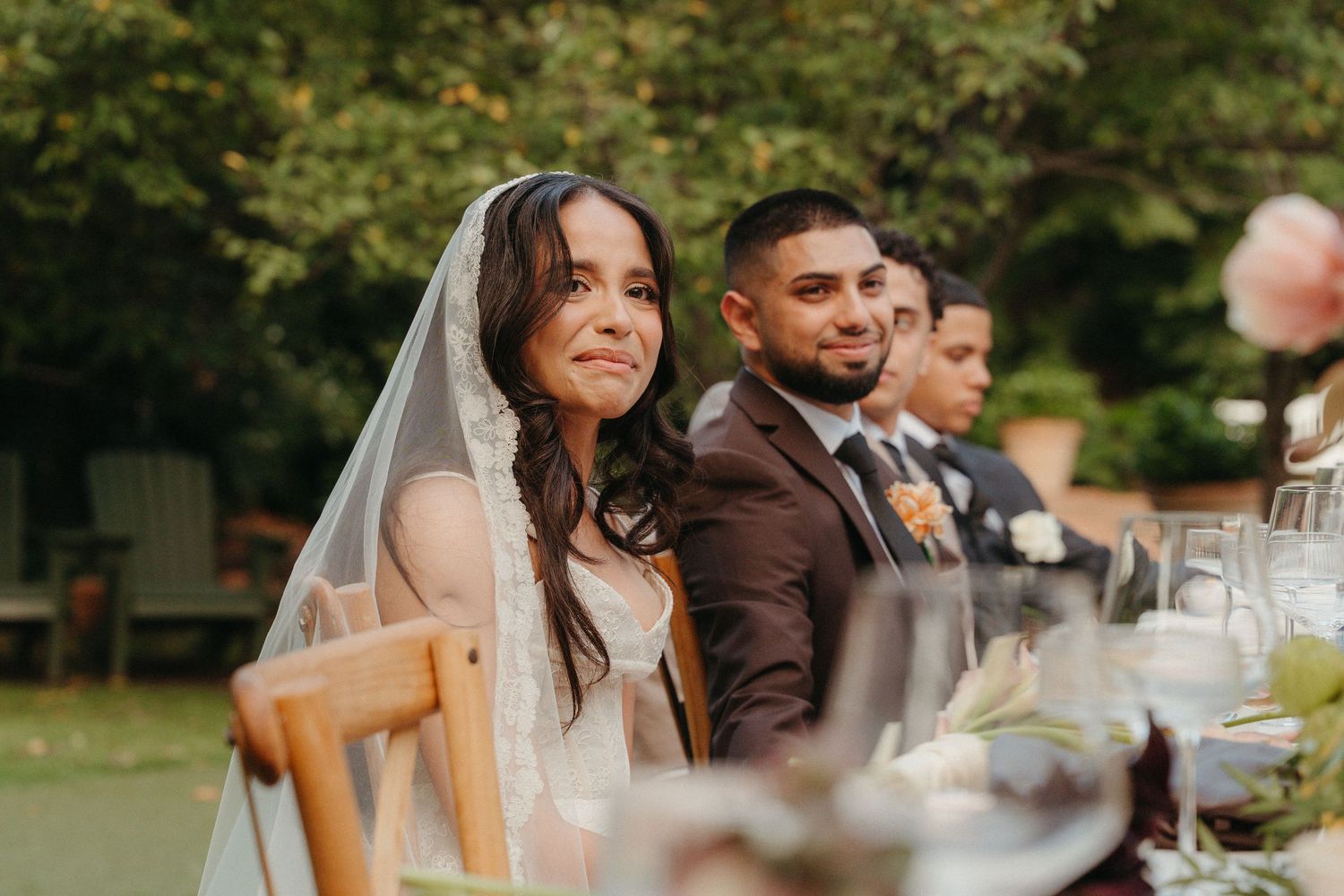 Bride in veil and groom in suit smile at outdoor wedding reception table surrounded by greenery.
