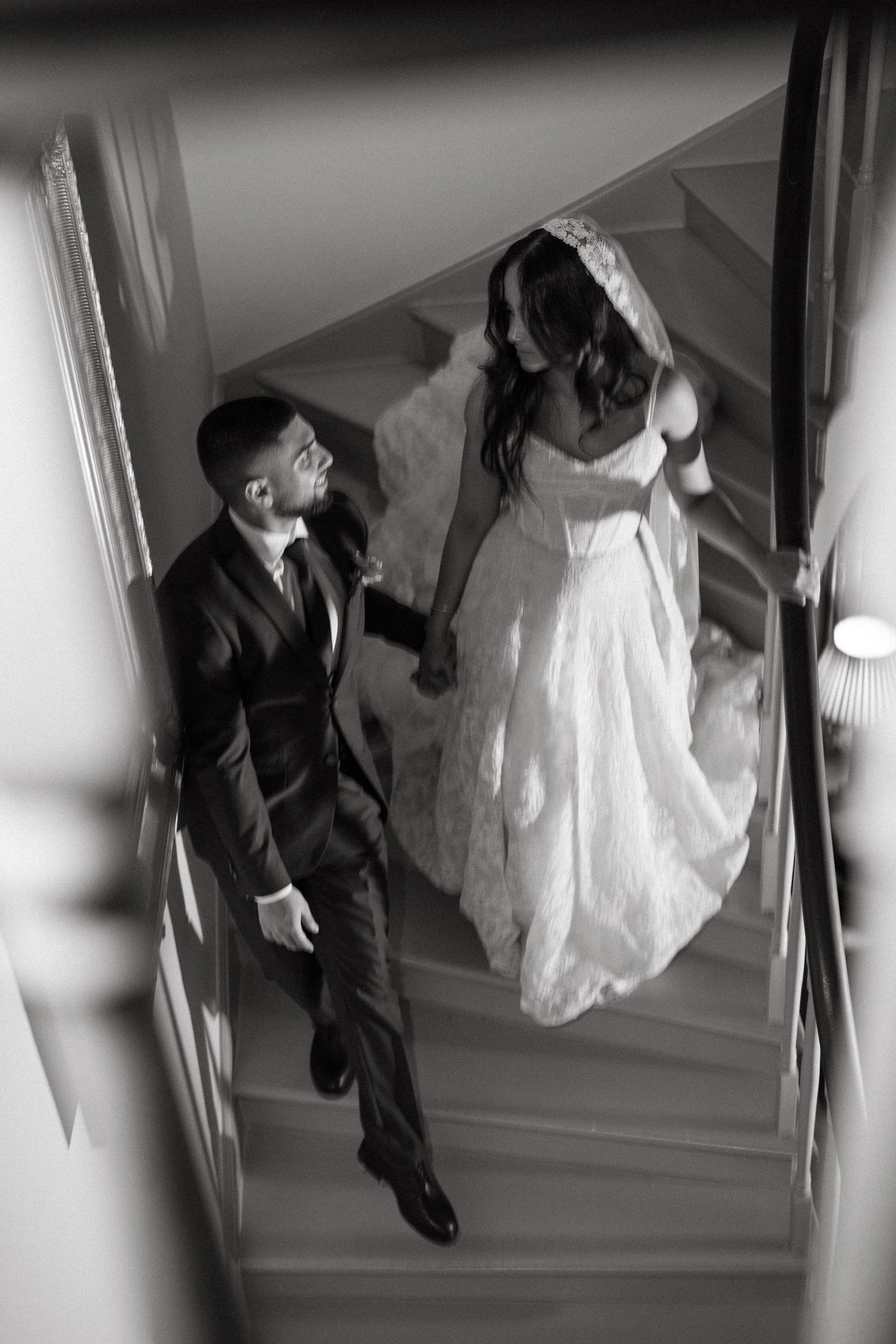 Black and white aerial photo of bride and groom holding hands walking up elegant staircase on wedding day.