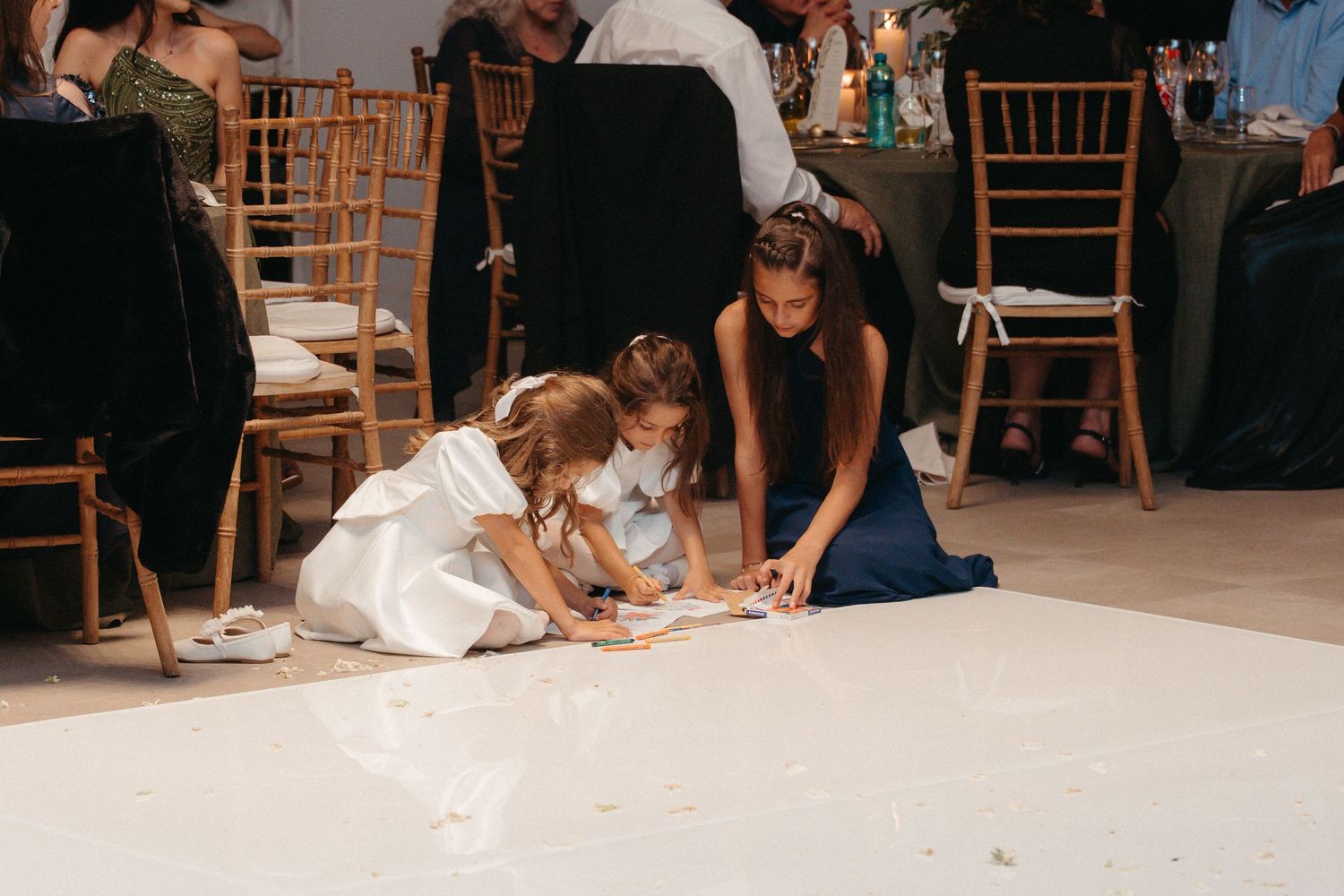 Three girls in formal dresses playing on a white dance floor at an elegant wedding reception.