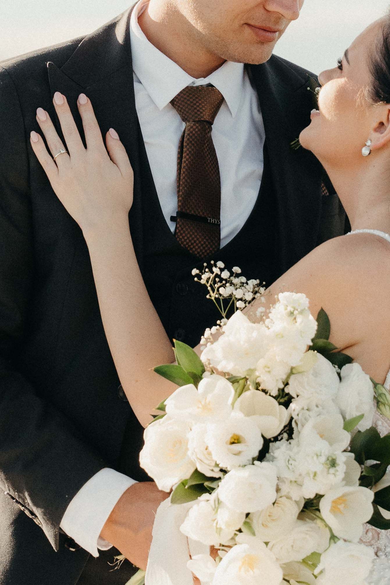 A bride holding a white floral bouquet gazes up at her groom in a black suit with brown tie on their wedding day.