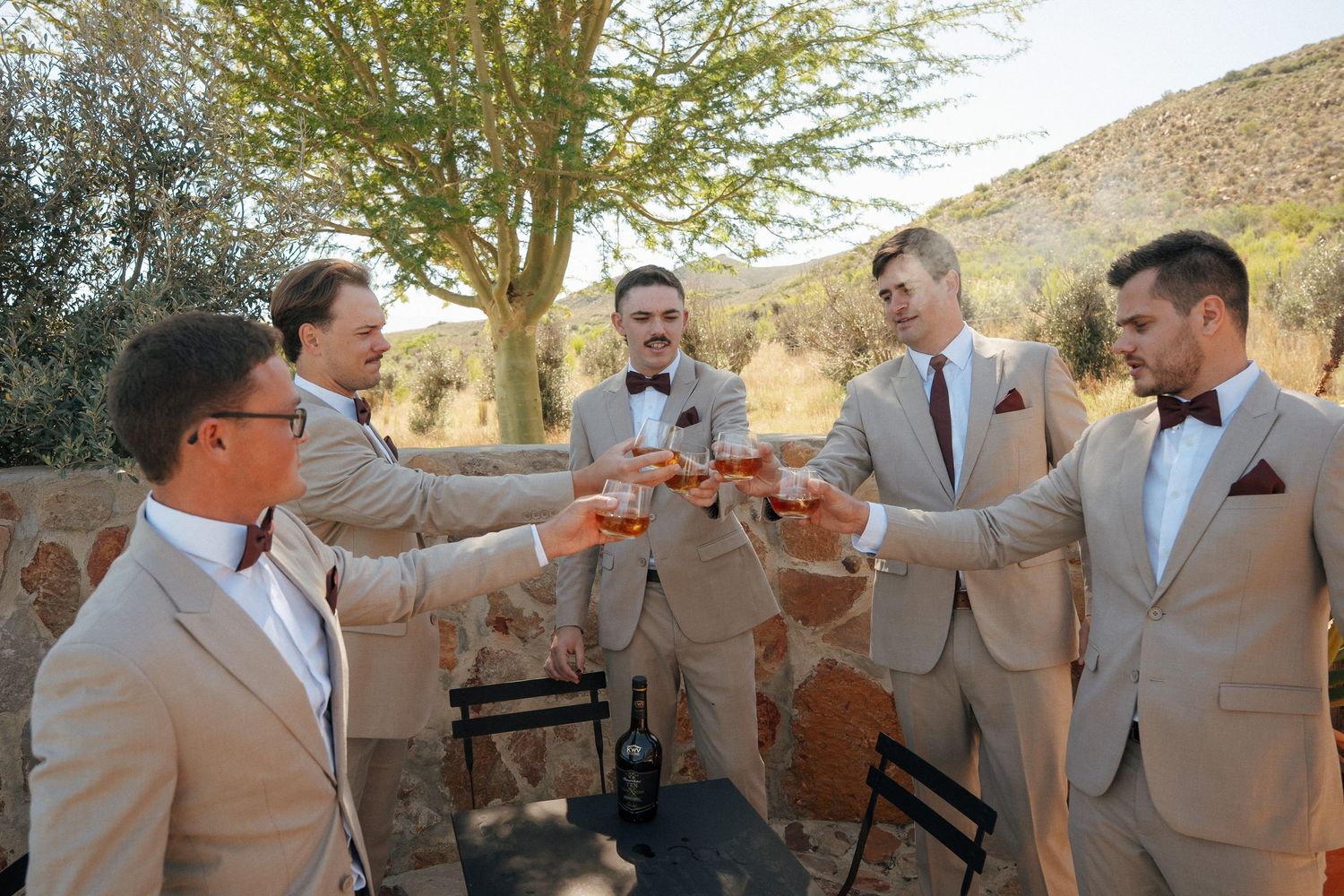 Five groomsmen in tan suits and bow ties raise glasses in a toast outdoors near a stone wall with Cape Winelands hills in the background.