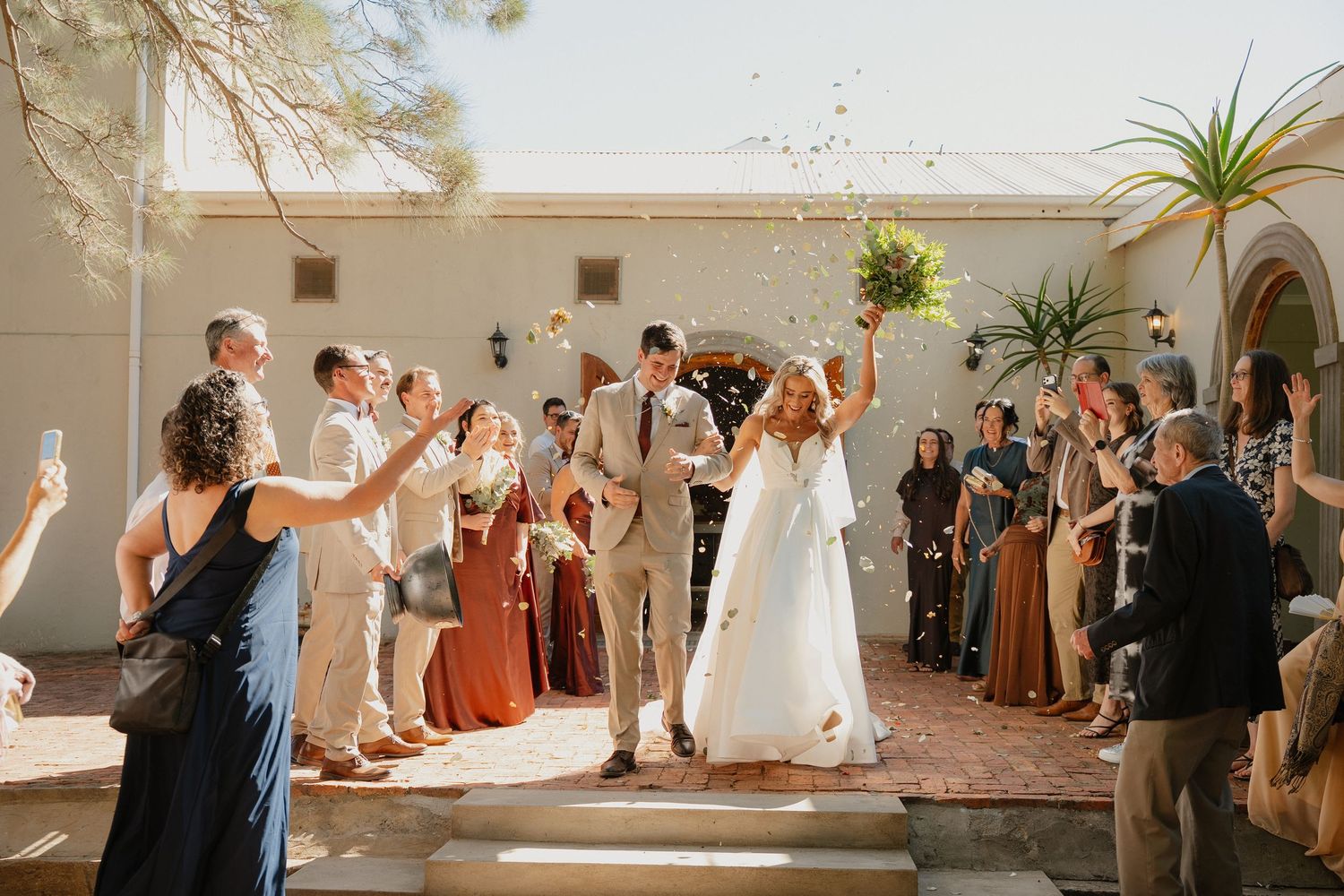 Bride and groom exit wedding ceremony as guests toss confetti outdoors at a sunny courtyard venue.