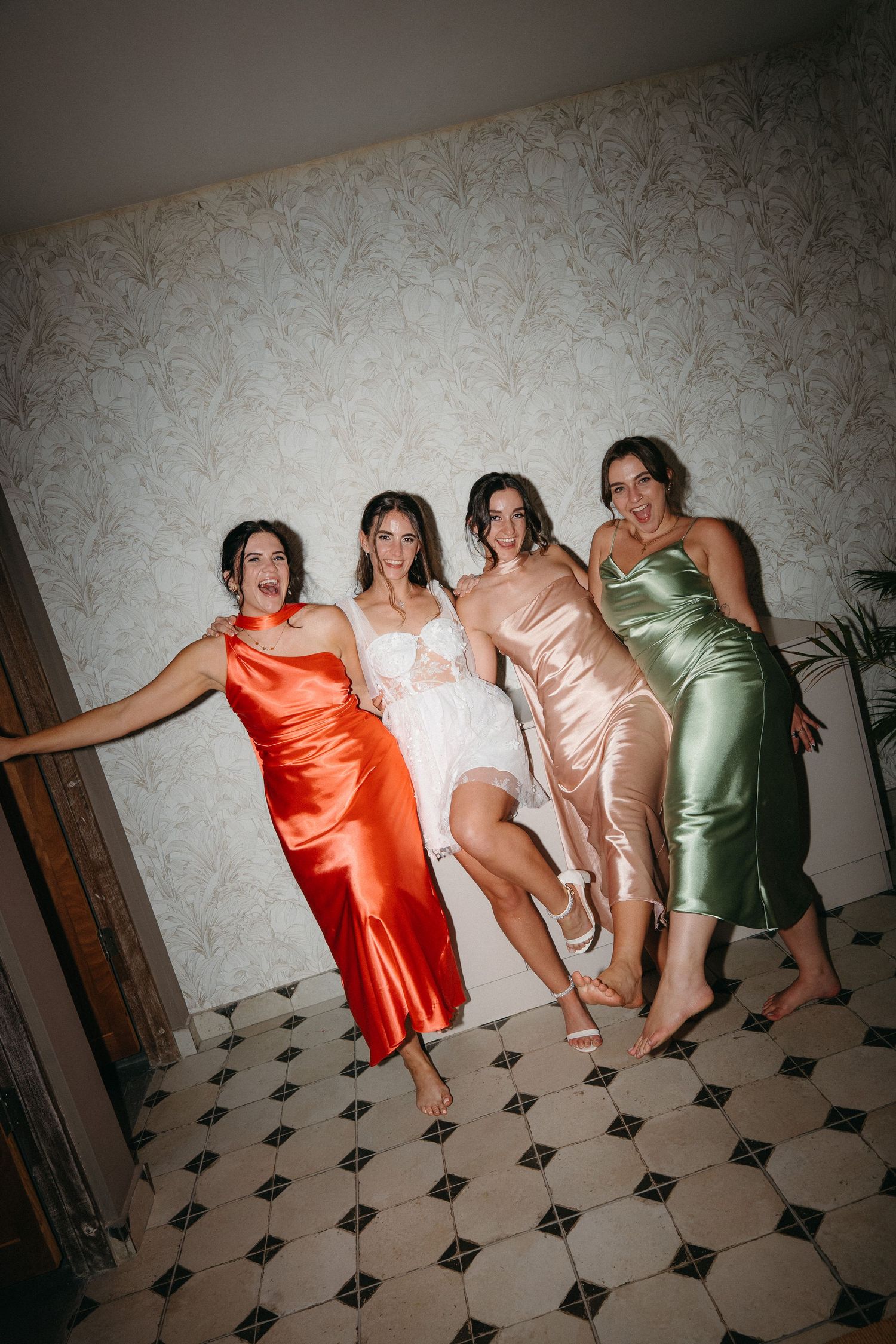 Bride and bridesmaids in satin dresses pose playfully against floral wallpaper on a black and white tiled floor.