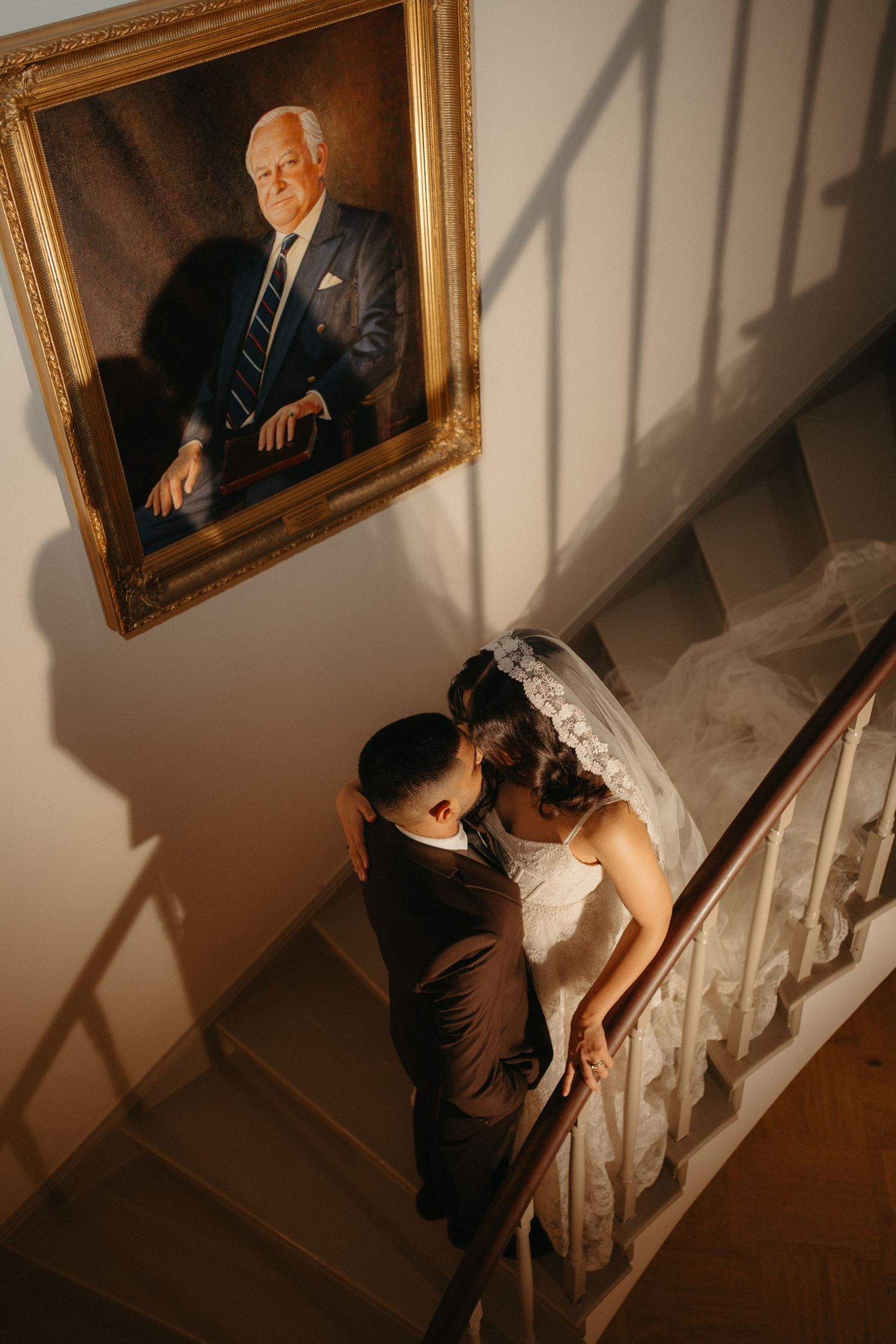 Bride and groom share a romantic moment on elegant staircase beneath a classic portrait painting in warm golden light.
