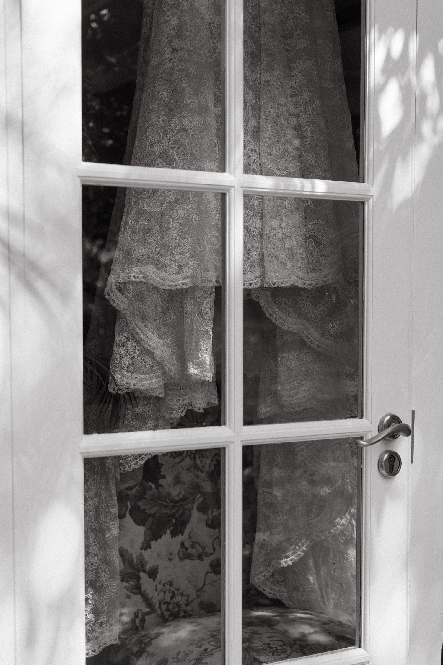Black and white photo of lace wedding dress hanging behind white French door with glass panes.
