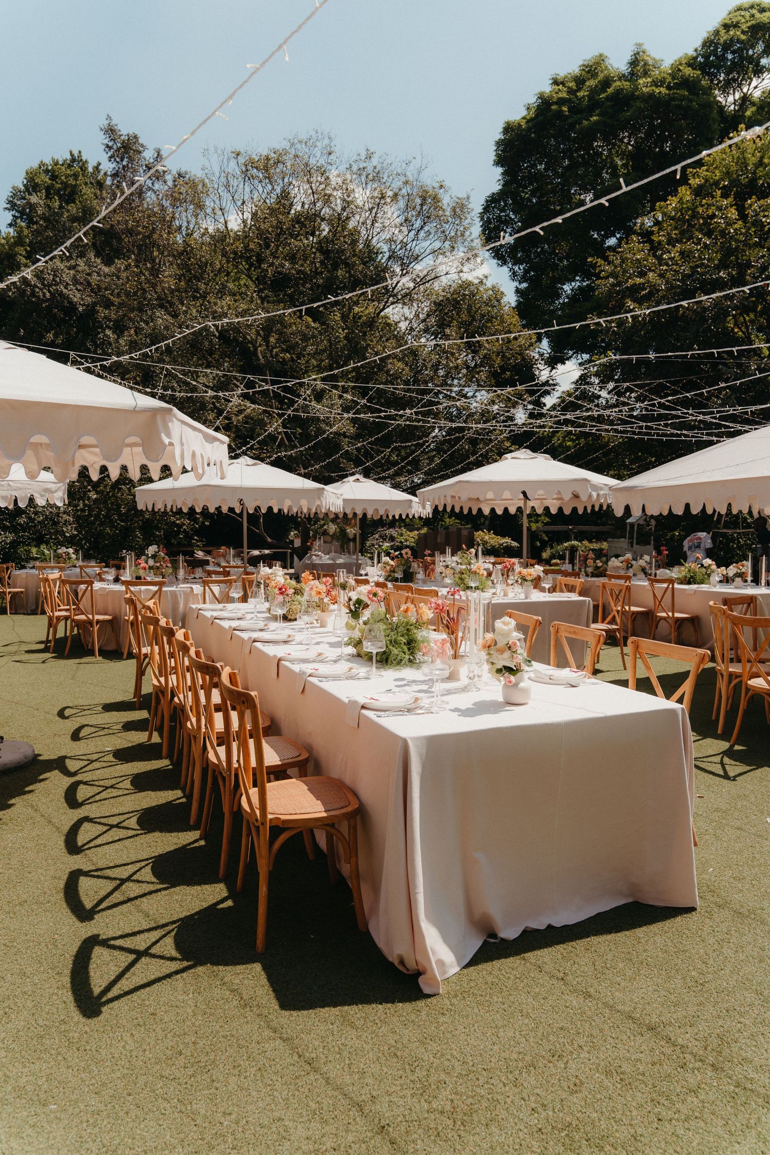 Elegant outdoor wedding reception with long white-draped tables, wooden chairs, string lights, and floral centerpieces on green lawn.