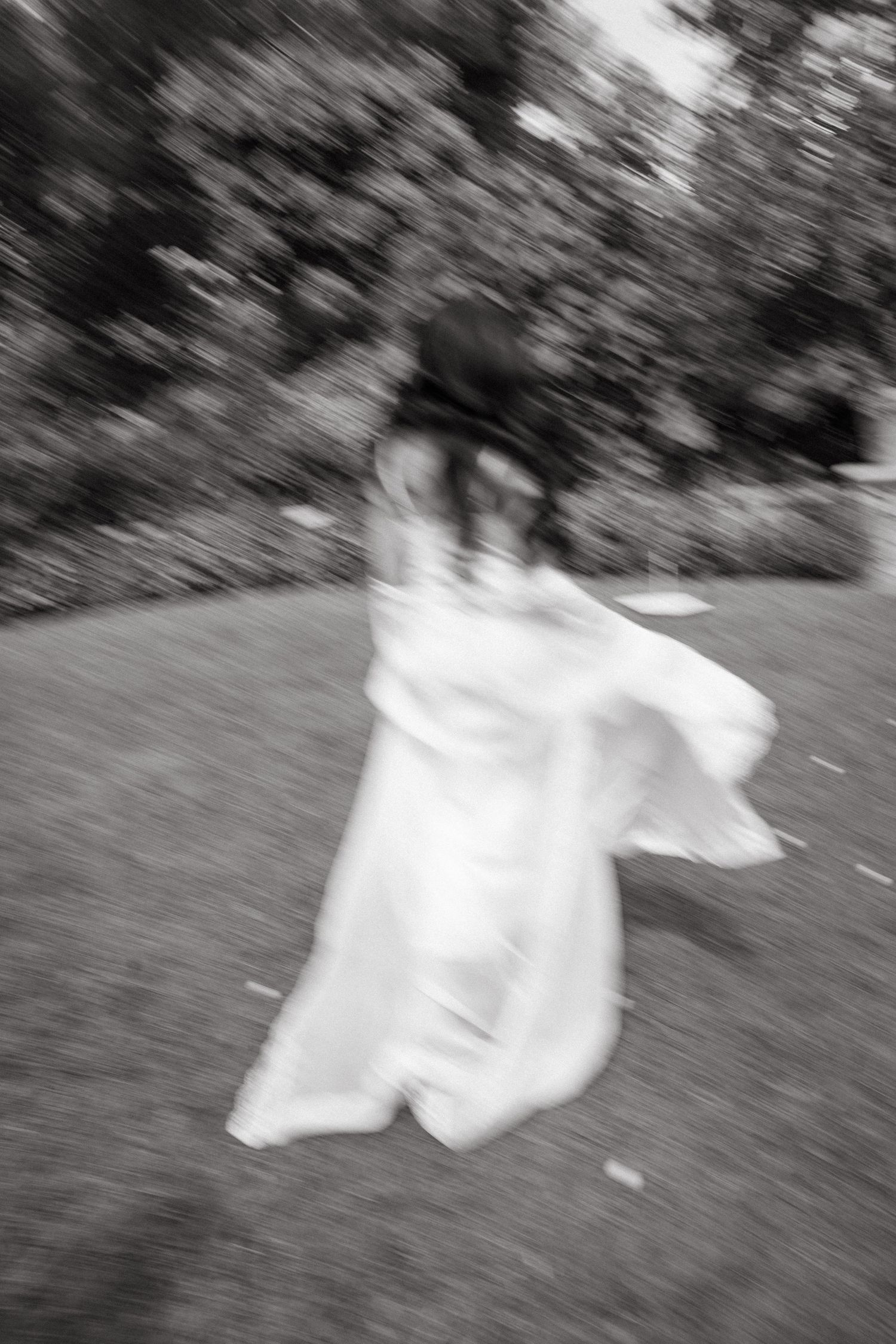 Blurred black and white photo of a bride in a flowing white gown spinning outdoors on grass near trees.