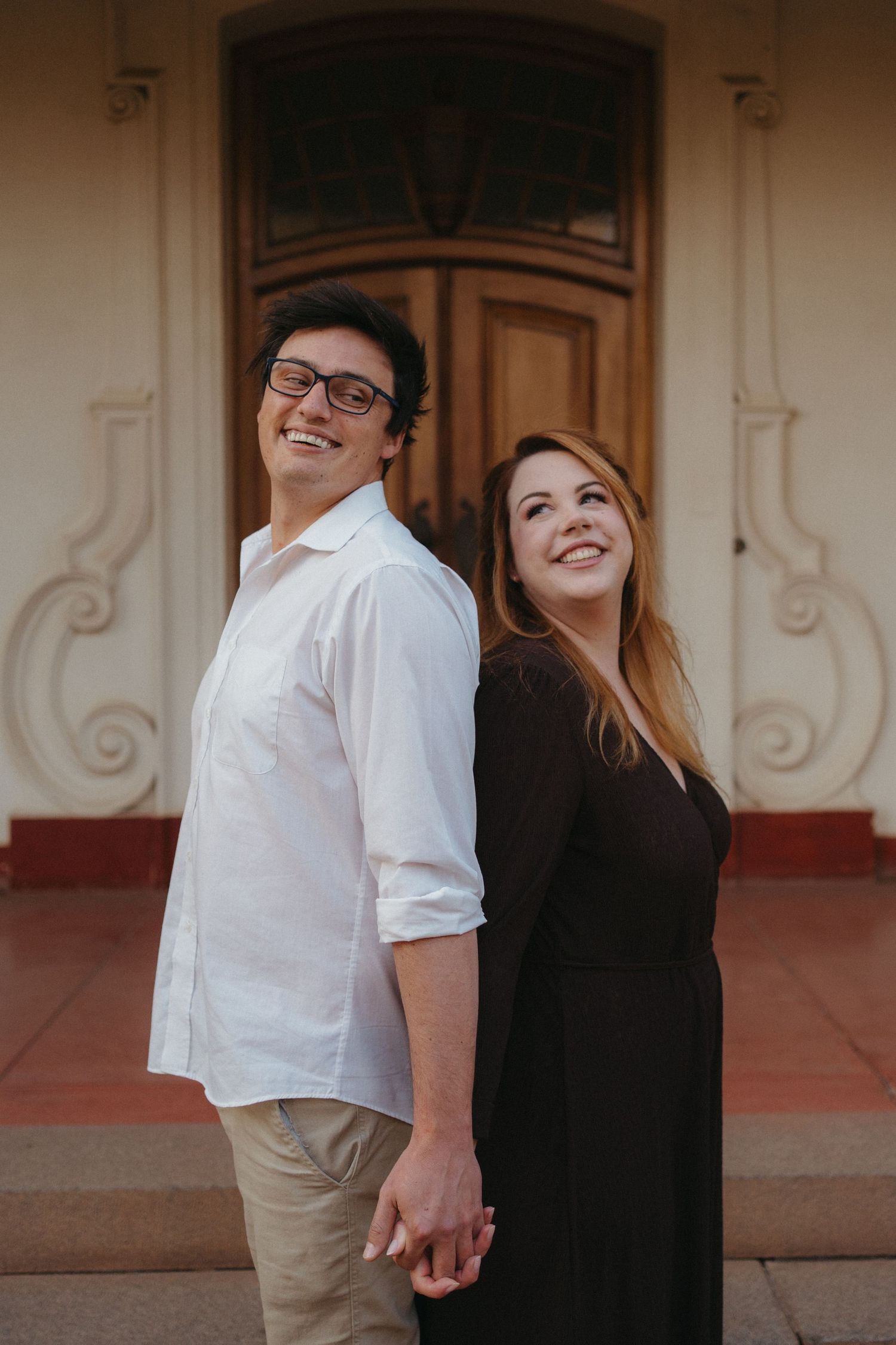 Smiling couple standing back to back holding hands in front of an ornate building entrance in Pretoria.