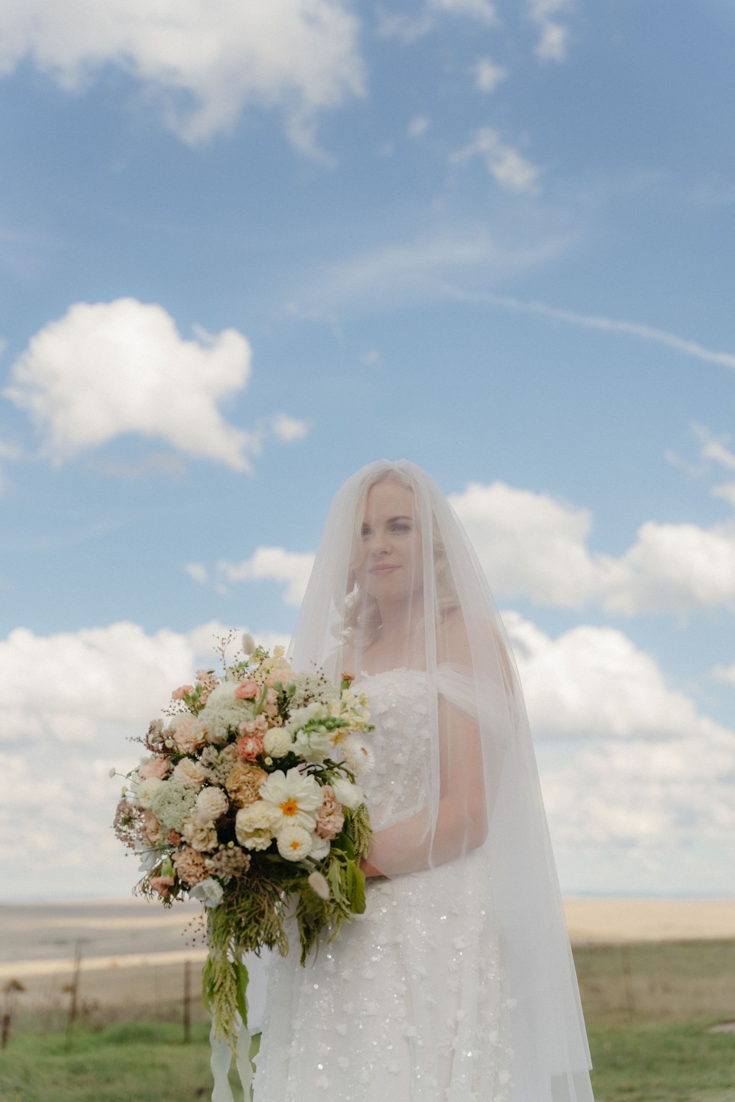 Bride in beaded gown holding floral bouquet under veil against blue Free State sky at Kasteelkop Guest Farm