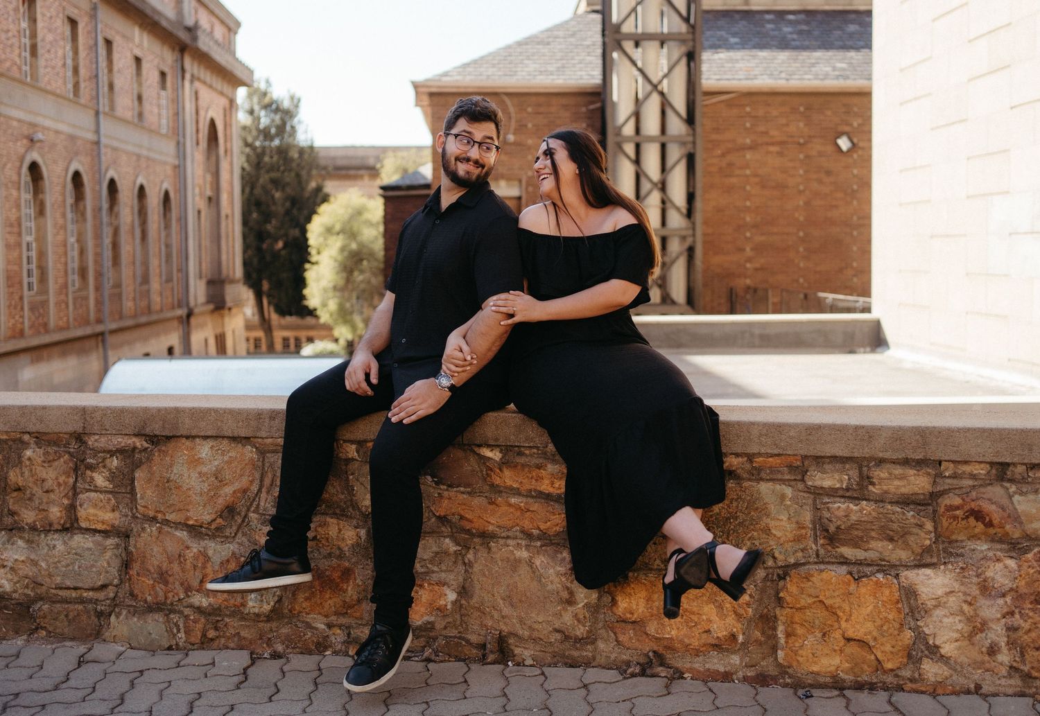 A couple sitting on a brick wall outside historic buildings in casual attire.
