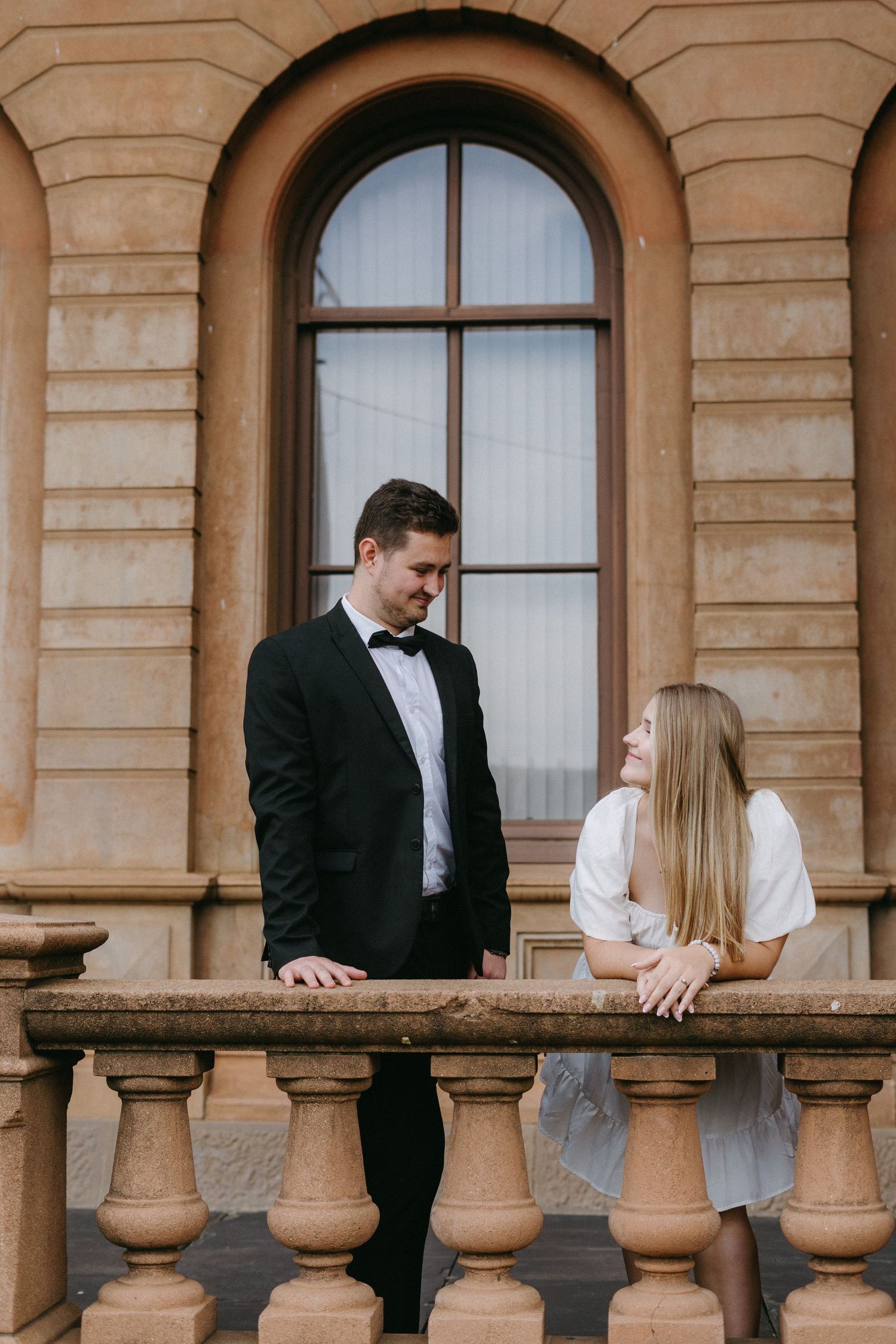 Couple in formal wear standing at ornate stone balustrade of historic building with arched windows.