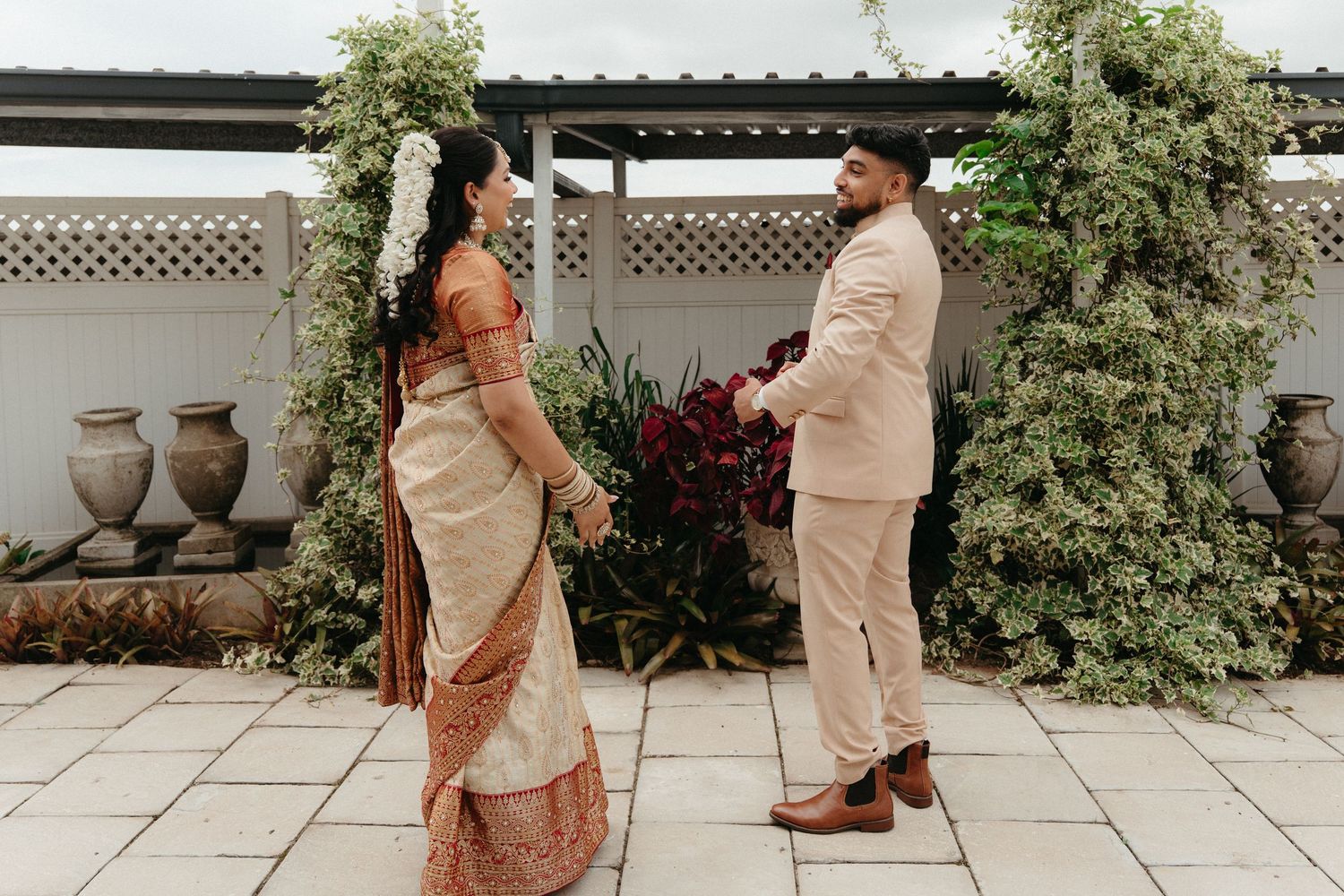 Two people pose together in matching beige and brown outfits on a white stone patio with greenery and pottery.