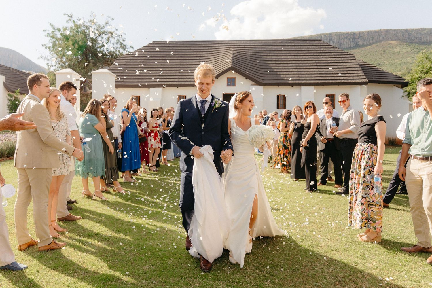 Wedding guests celebrate by tossing confetti at a sunny outdoor ceremony with mountains in the background.