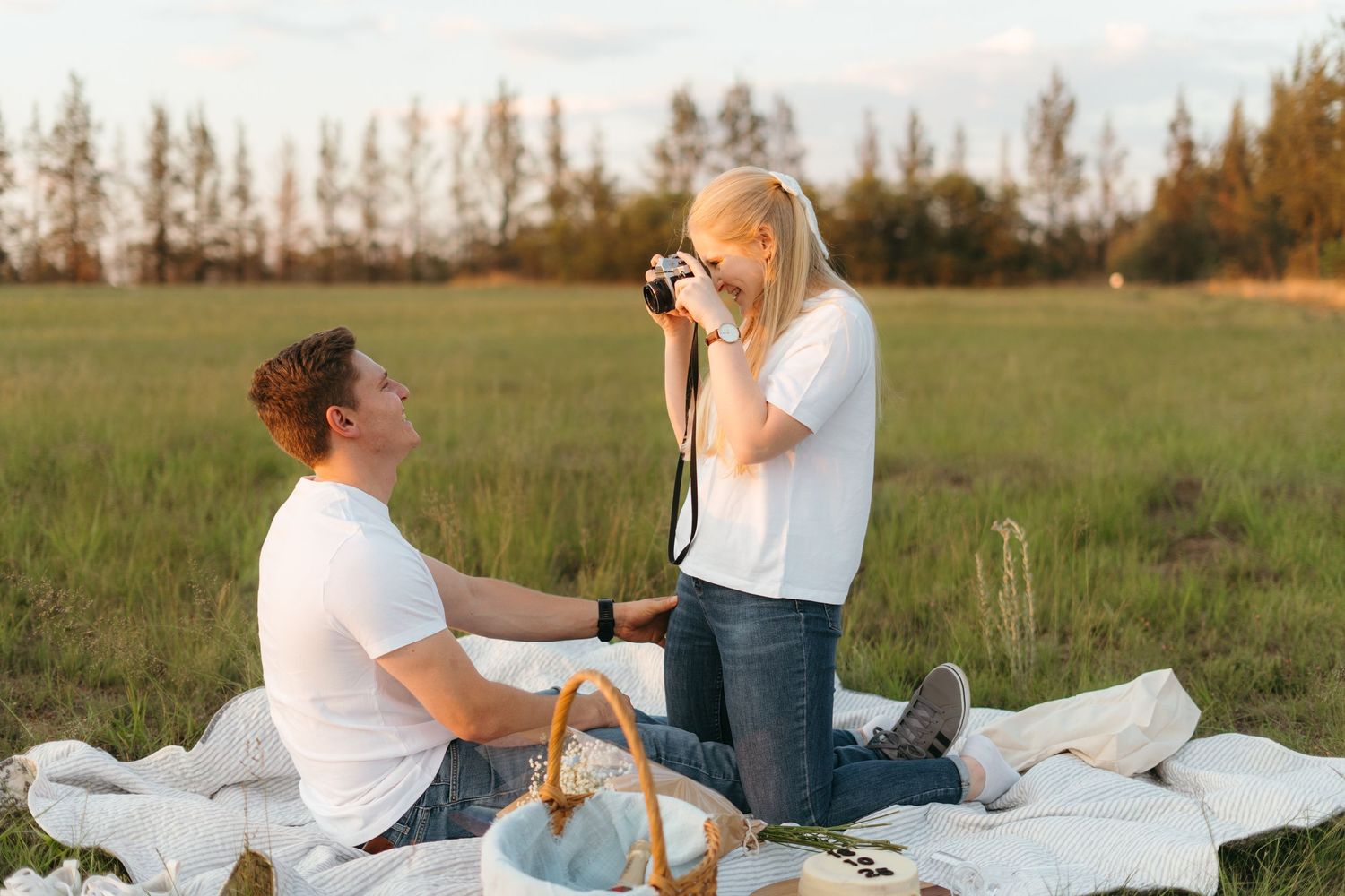 A couple enjoys a romantic picnic on a blanket in a grassy field during golden hour while taking photographs.