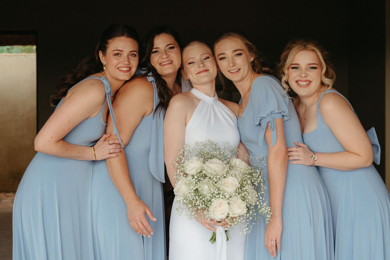 Bridesmaids in light blue dresses pose together with the bride holding a white floral bouquet.